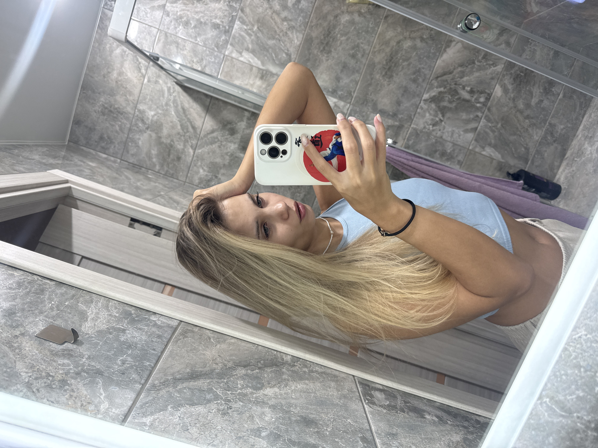 A young woman is captured in a mirror selfie within a tiled bathroom, oriented diagonally. She has long, blonde hair and is wearing a light blue ribbed top and light-colored shorts. Her head is slightly tilted as she looks towards her smartphone, which she holds up to take the photo.
The phone has a distinctive white case featuring a red circular emblem with a Japanese-style illustration of two figures, possibly engaged in martial arts like judo or sumo, and visible Japanese characters. One character within the red circle appears to be "柔" (ju) and other characters are also present.
The background shows a contemporary bathroom with grey, marbled tiles on both the floor and walls. A glass shower enclosure with chrome fixtures is visible in the upper part of the reflection. Inside the shower area, a purple towel or piece of fabric rests on the tiled floor next to a dark-colored bottle, possibly a personal care product. A small, metallic, irregularly shaped object lies on the tiled floor in the lower left foreground. The scene depicts an indoor setting, likely in Chaikovsky, Russia, as suggested by the context.