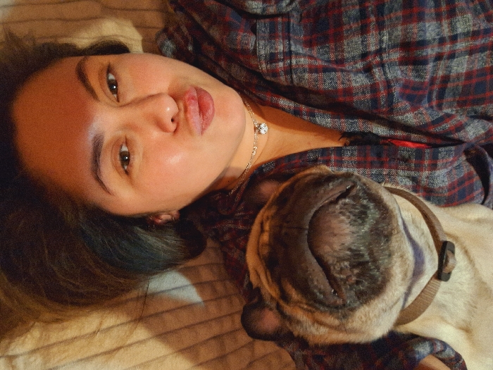 A woman lies on a striped surface, possibly a bed. She wears a plaid shirt and a necklace with a sparkly pendant. Beside her is a sleeping pug, its face upside down and close to the camera. The pug has a brown collar. The woman looks at the camera with slightly parted lips. Her hair is dark, and she has light skin.