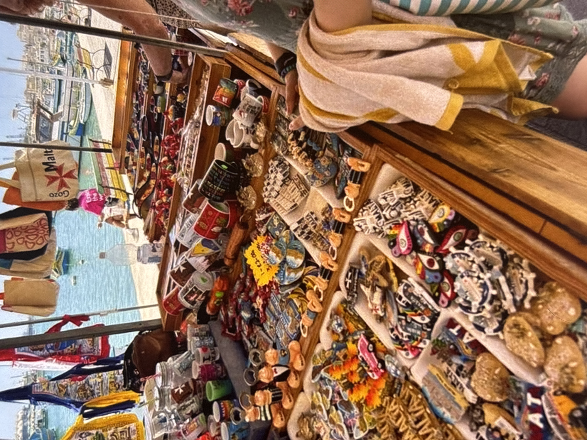 The image depicts an outdoor market stall overflowing with various souvenirs and trinkets. The stall is set up in a harbor or waterfront area, with a body of water visible in the background, dotted with boats. The stall itself is constructed from wood and metal, with shelves and trays displaying a wide array of items.
Dominating the foreground are numerous small souvenirs, many of which appear to be magnets or decorative items. These include representations of landmarks, local symbols, and small figurines. Some items are arranged in trays, while others are displayed more loosely. Notably, there are several mugs or cups with printed designs, and some items are colorful and playful, such as small figures of people in formal wear. A yellow price tag is also visible, indicating that transactions are taking place.
Behind the merchandise, glimpses of people can be seen. A person in a bright pink shirt is sitting on a bench in the background. Another person, partially visible on the right side of the frame, appears to be a vendor or customer, with their hand reaching towards the merchandise. They are wearing a patterned dress or shirt and have a towel draped over their arm. A bag with the word "Malta" is hanging prominently from the stall's structure.
The overall scene suggests a vibrant tourist area where local crafts and souvenirs are being sold to visitors. The lighting indicates it is daytime, and the presence of boats and the waterfront suggests a coastal or port city. While the prompt mentions Orihuela, Spain, the visual cues in the image, such as the "Malta" branding and the style of boats and architecture in the distance, strongly suggest the location is actually Malta, not Orihuela. The items for sale are typical of tourist markets in Malta.