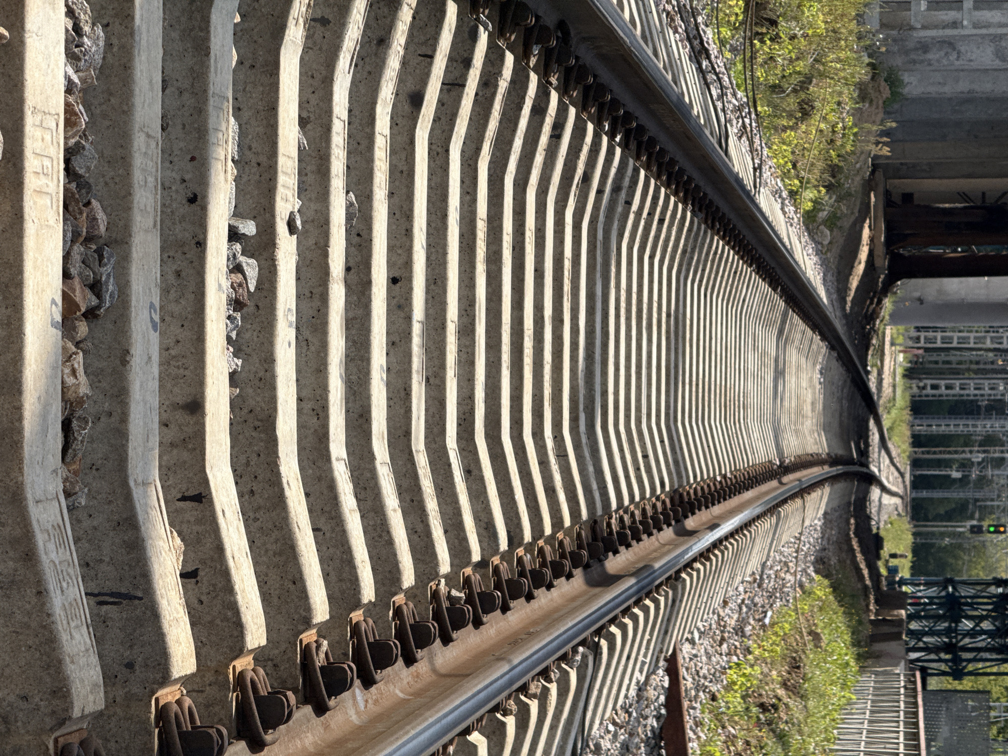 A close-up, low-angle perspective shot captures a railway track in Khimki, Russia, on a sunny day. The image focuses on the concrete railway ties, which are laid out in a pattern of alternating light and shadow due to the sunlight. Metal clips secure the dark steel rails to the concrete ties. Small stones and gravel are scattered between the ties, with some areas appearing more weathered and dusty than others. In the background, the railway track curves towards the left, leading to a junction where another set of tracks diverges. Above the tracks, a green traffic light is visible, indicating it is currently safe to proceed. The surrounding environment includes green foliage on the left and right sides of the tracks, as well as a metal fence and structural elements that suggest the presence of infrastructure. The scene appears to be located in an outdoor industrial or transportation area.