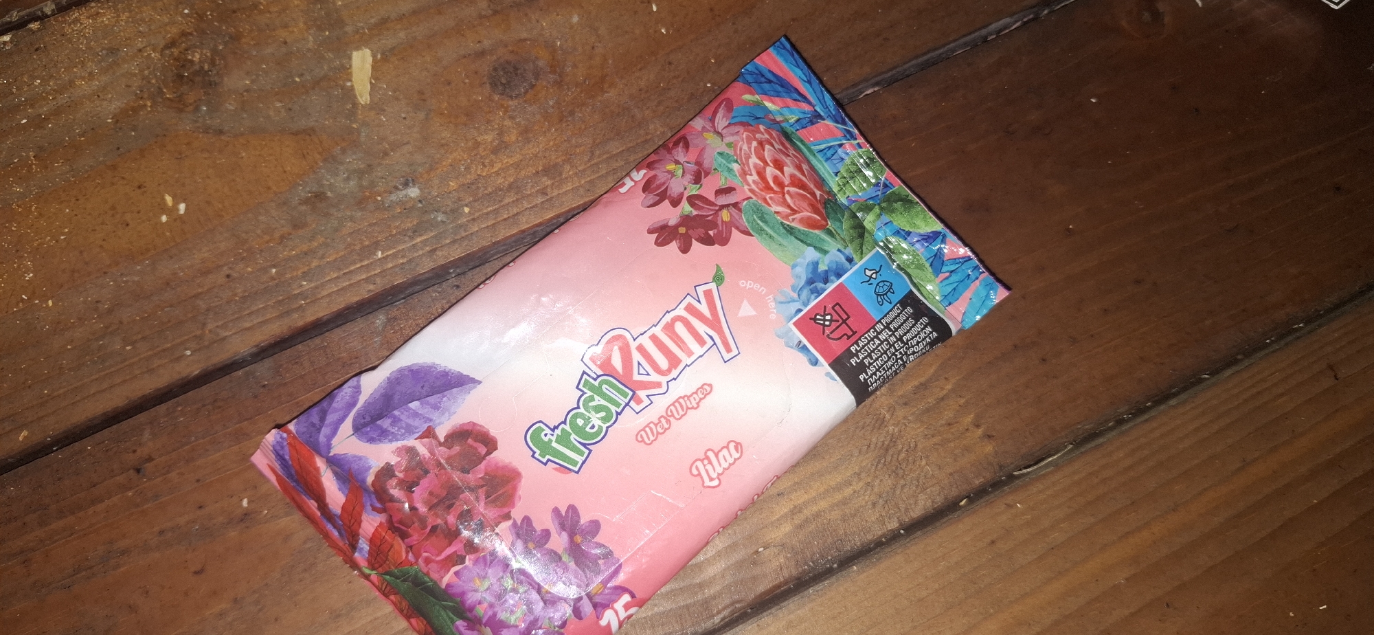 A pink package of "fresh Runy Wet Wipes" with a floral design is lying diagonally on a wooden surface. The package has text indicating "Lilac" and "Wet Wipes." There are small symbols on the lower right corner of the package. The wooden surface appears to be made of planks and has some debris on it. The scene is a close-up shot, with the focus on the product packaging and the texture of the wood. There is no indication of the specific location within the city of Shumen, Bulgaria.