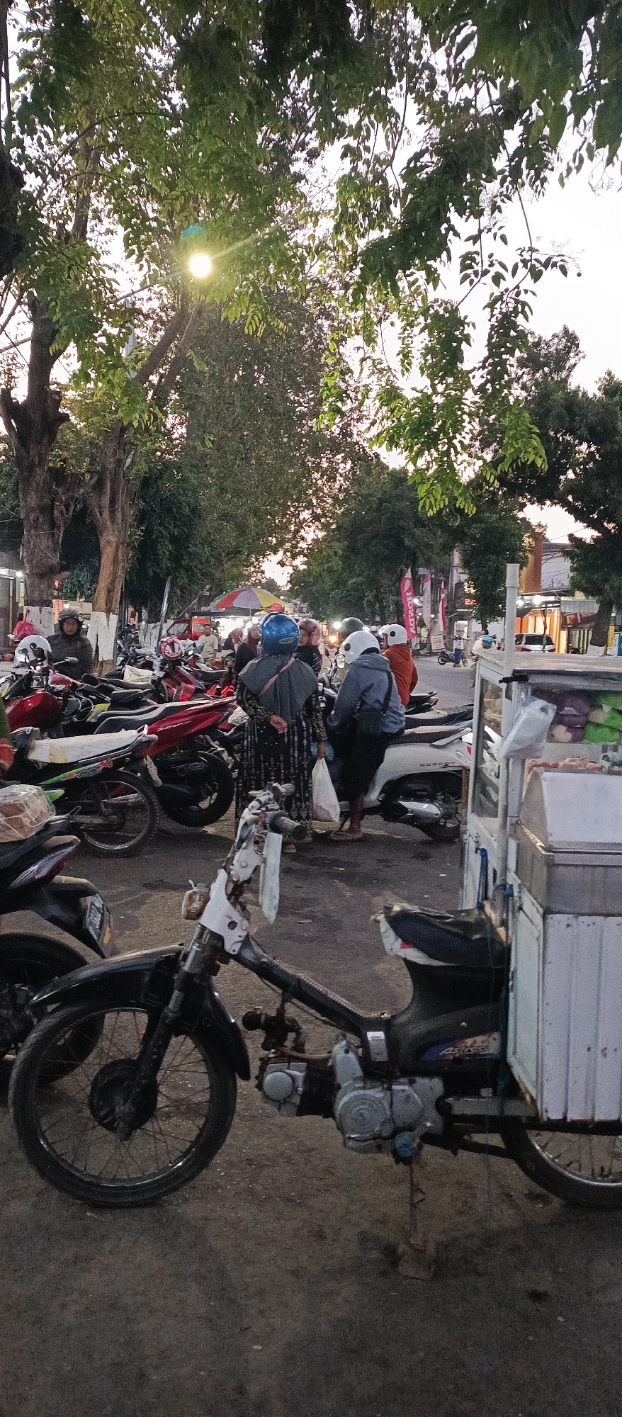 This is an outdoor, eye-level shot of a street scene at dusk in Ardirejo, Indonesia. In the foreground, a black motorcycle is parked, with its engine and wheels visible. Behind it, a white food cart is parked, displaying various food items on its shelves. A group of people, some wearing helmets and traditional Indonesian clothing, are gathered around several motorcycles, some of which appear to be parked in a market area. In the background, there are trees, buildings, and streetlights, suggesting a commercial or residential area. The sky is a light orange, indicating the sun is setting.