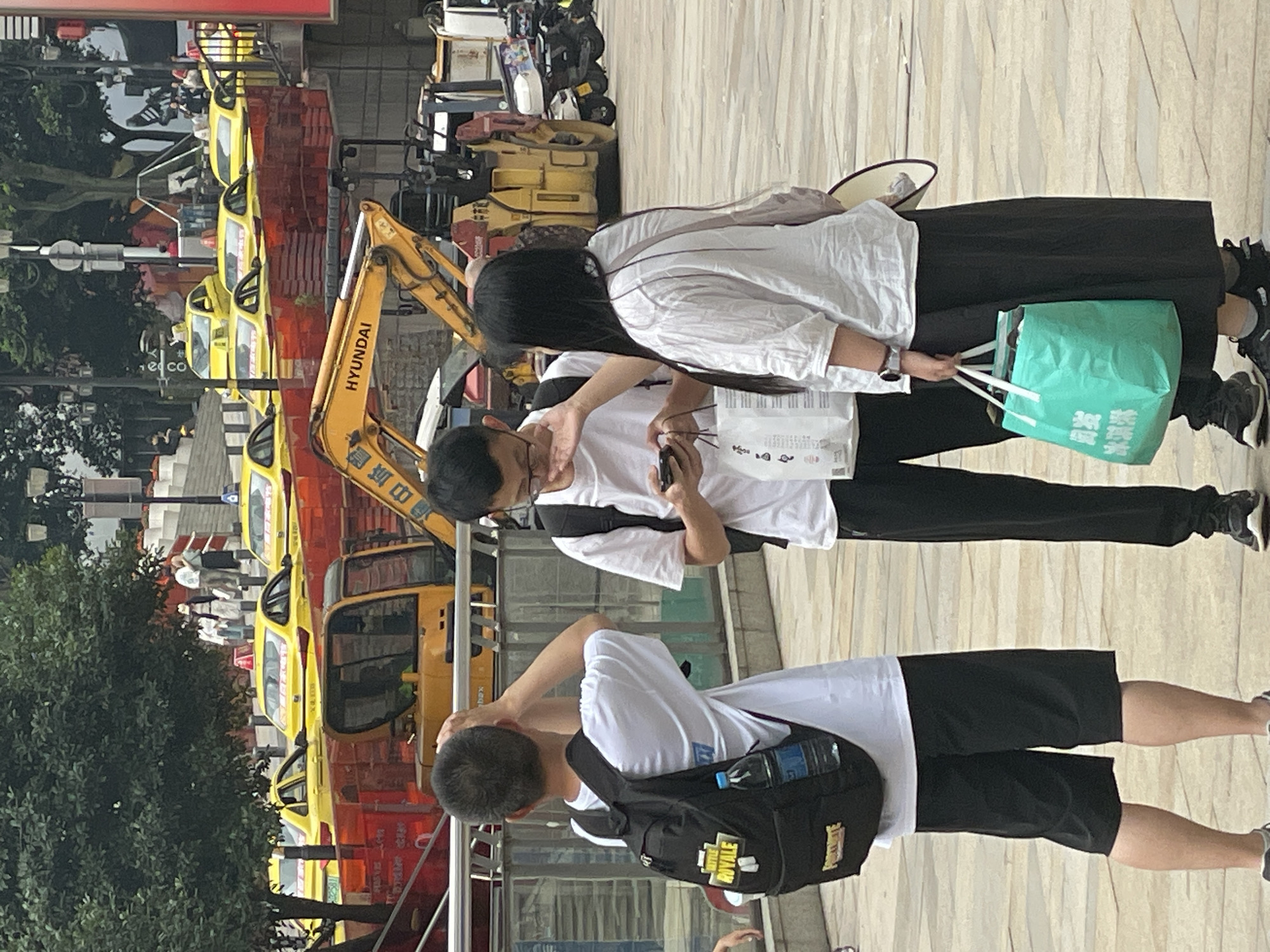 Three individuals are walking on a sidewalk. One person is wearing a white shirt and black shorts with a black backpack. Another is wearing a white shirt and black pants. The third individual is wearing a white shirt and black pants and carrying a green shopping bag. There is a row of yellow vehicles, a construction digger, and a tree visible in the background. The location appears to be Yuzhong District, China.