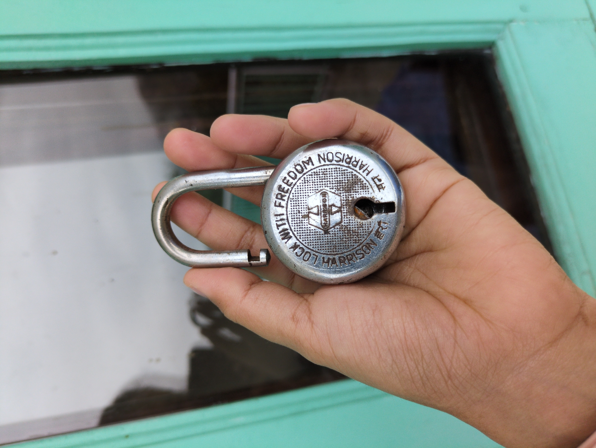 A hand holds a silver padlock with the shackle open. The padlock has text embossed around its circular body, which reads: "LOCK WITH FREEDOM HARRISON INDIA". Below this, in smaller text, it says "सन HARRISON" which translates to "Sun Harrison". The padlock is centrally positioned within the frame. The hand holding the padlock appears to be of Indian descent, with visible palm lines. The background consists of a blurred green-painted window frame and reflective glass, suggesting an indoor or outdoor setting with a reflective surface. The overall scene is brightly lit, with a slight overexposure.