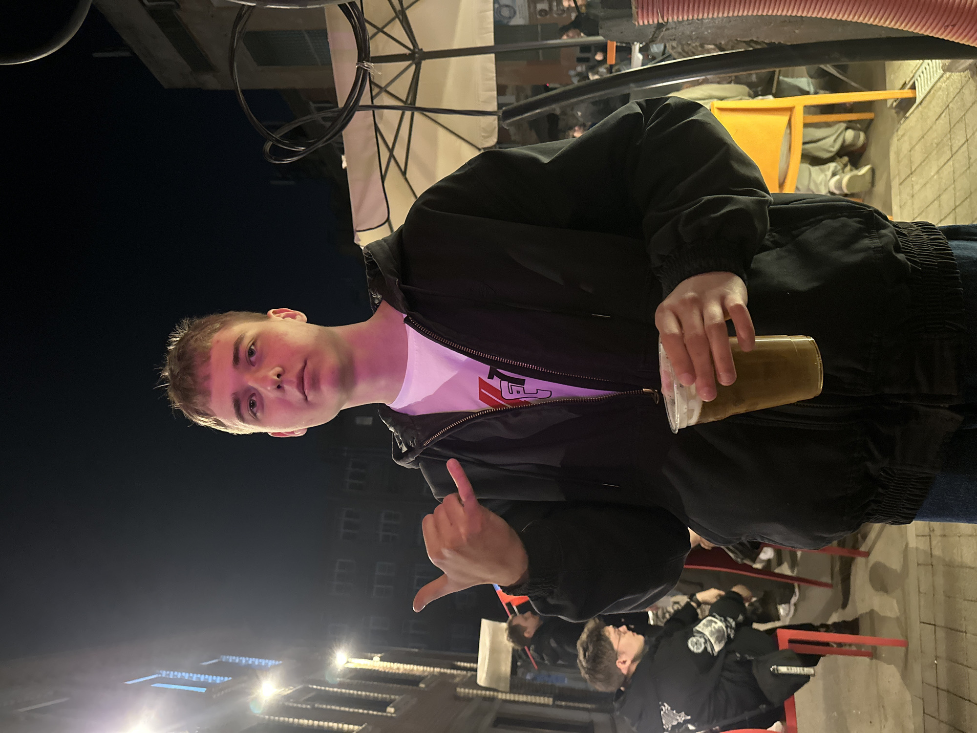 The photo depicts a young man in a black jacket and white t-shirt holding a beverage. He is gesturing with his hand. The setting appears to be outdoors at night, possibly in Krasnoyarsk, Russia, based on visual cues like building facades and street lighting. Other individuals are visible in the background, seated at tables. The photo includes outdoor furniture like chairs and tables and an umbrella structure.