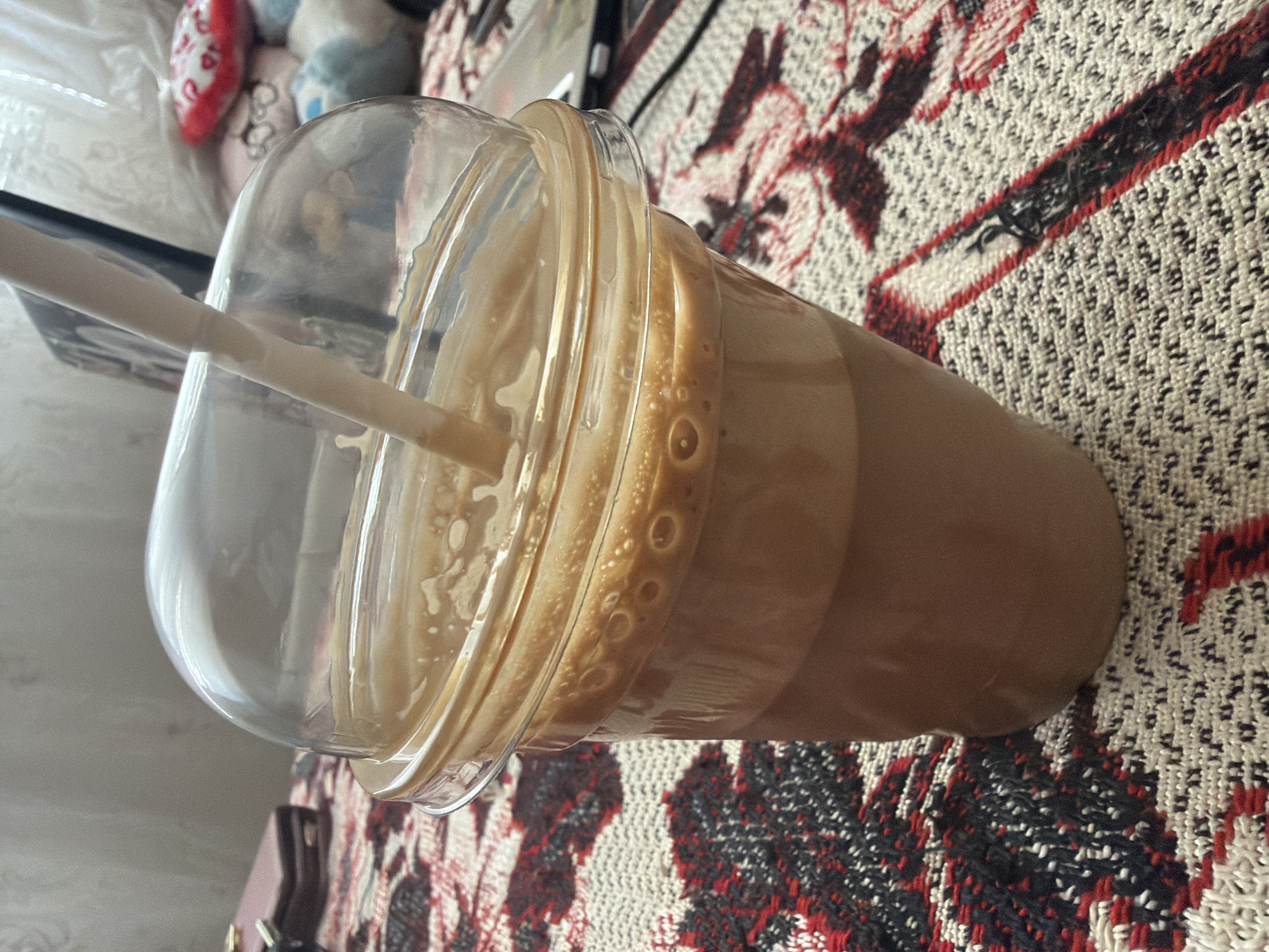 A tilted, close-up shot shows a plastic cup filled with a beige liquid, likely a coffee-based drink, with a dome lid and a straw inserted. The cup sits on a textured fabric surface with a pattern of interwoven white, red, and black elements. The scene is softly lit, highlighting the bubbles and texture of the drink. The location is impossible to determine from the given image.