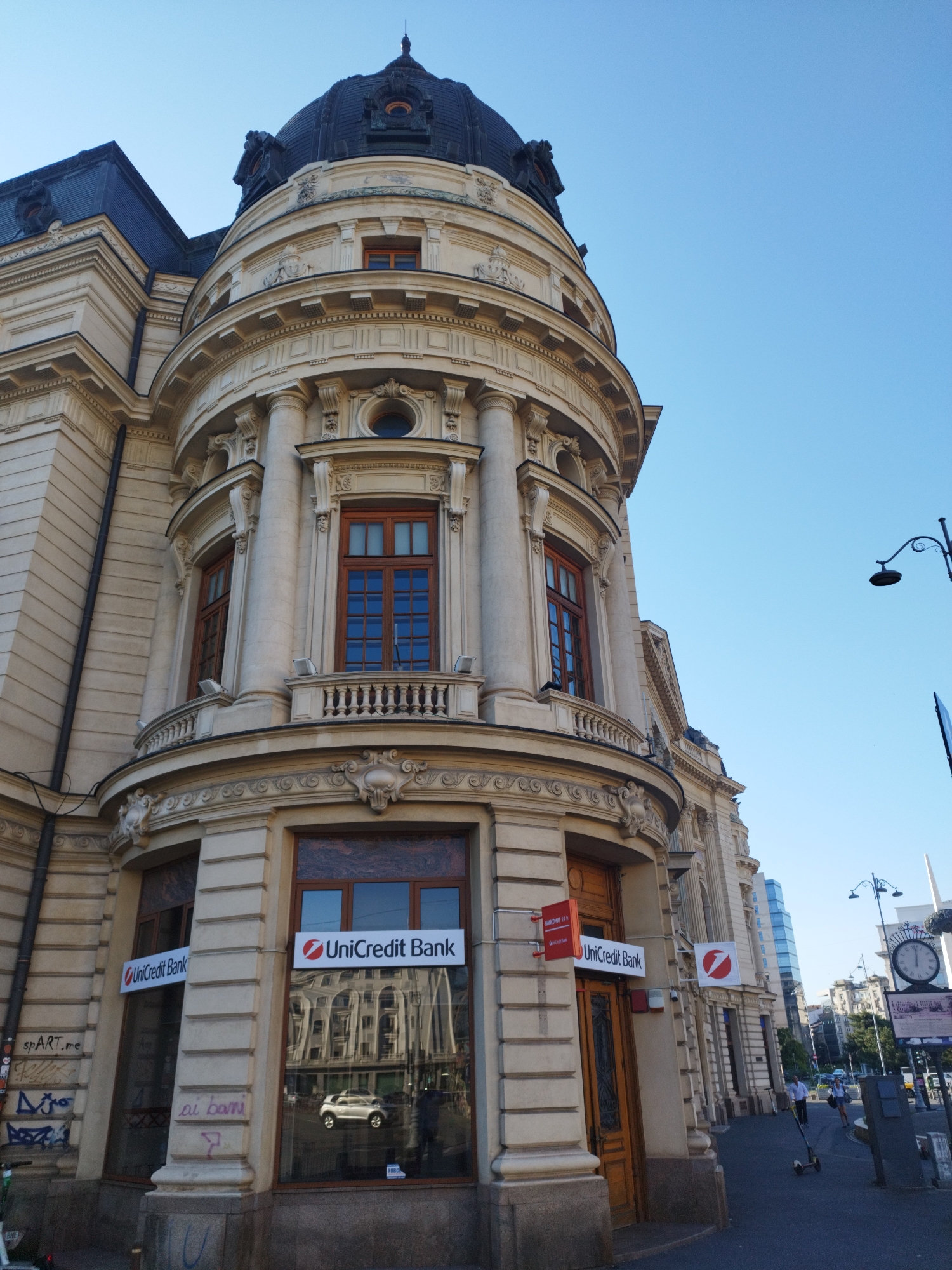 The image shows the exterior of a grand, ornate building with a dark dome, likely in the city of Bucharest, Romania. The building features classical architectural elements such as columns, balconies with decorative railings, and large windows with wooden frames. Several "UniCredit Bank" signs are visible on the ground floor, indicating it is a bank branch. One window displays a reflection of a white car and other buildings. In the background, a street scene unfolds with people on scooters, a lamppost, a public clock, and modern buildings. The sky is clear and blue. The lower part of the building's facade has graffiti.