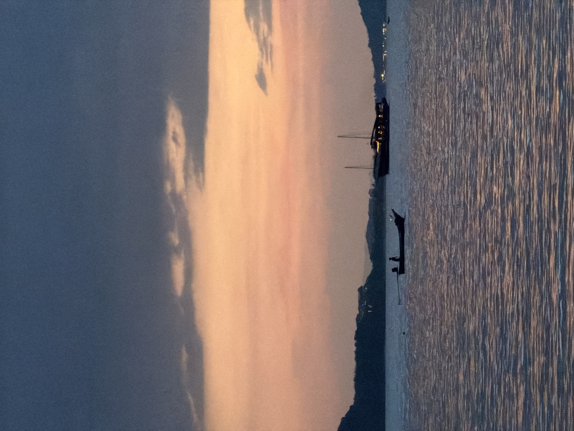 A vertical, silhouetted photograph captures a serene evening seascape at dusk, likely in Ko Samui District, Thailand. The sky is a gradient of dark gray clouds at the top, transitioning to a soft, warm peach and orange hue in the middle, with hints of lavender near the horizon. Below the sky, a dark, silhouetted landmass, likely an island or coastline, stretches across the frame, with faint lights suggesting habitation.
The foreground is dominated by the shimmering surface of the sea, reflecting the colors of the sky in elongated, horizontal patterns. Two boats are visible on the water. In the mid-ground, a narrow, traditional longtail boat is positioned centrally, with two indistinct human figures silhouetted within it. Further to the right, a larger, dark-hulled boat with visible lit windows and two masts floats on the water, also silhouetted against the colorful sky. The overall atmosphere is tranquil and picturesque, evoking a sense of calm at the end of the day.