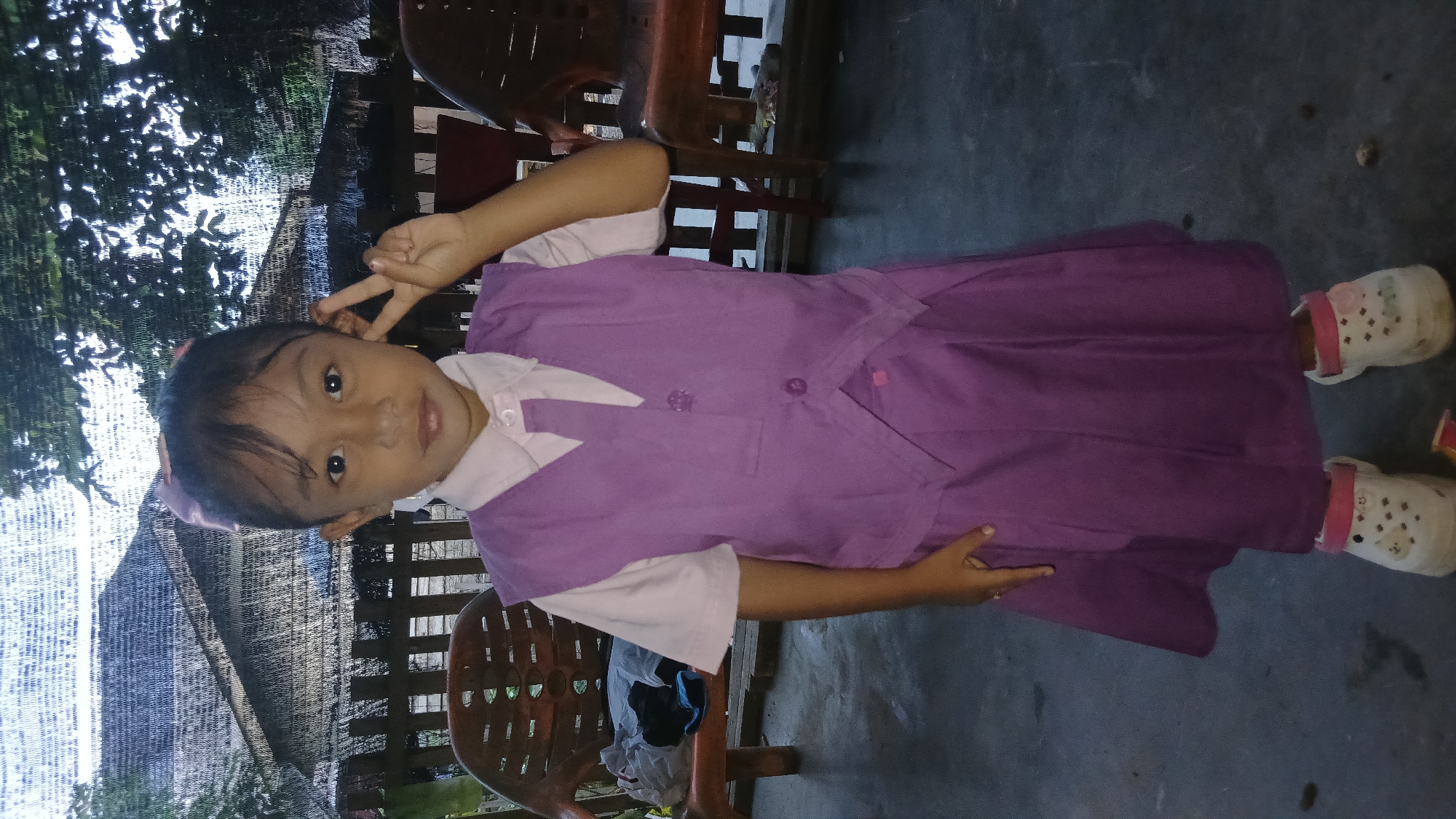 A young girl stands in a purple school uniform and white clogs on a paved surface, posing for a photo. She is wearing a light pink collared shirt with a purple vest and a matching purple skirt. The vest has two buttons and a decorative trim. Her left hand is raised to her face, with two fingers extended in a peace sign. She has dark hair pulled back with a pink bow. In the background, there are wooden chairs, a thatched roof structure, and some greenery. The scene appears to be outdoors, possibly in a yard or courtyard. The overall lighting suggests it is daytime, but possibly overcast or late in the day. The location is identified as Keudejungkagajah, Indonesia.