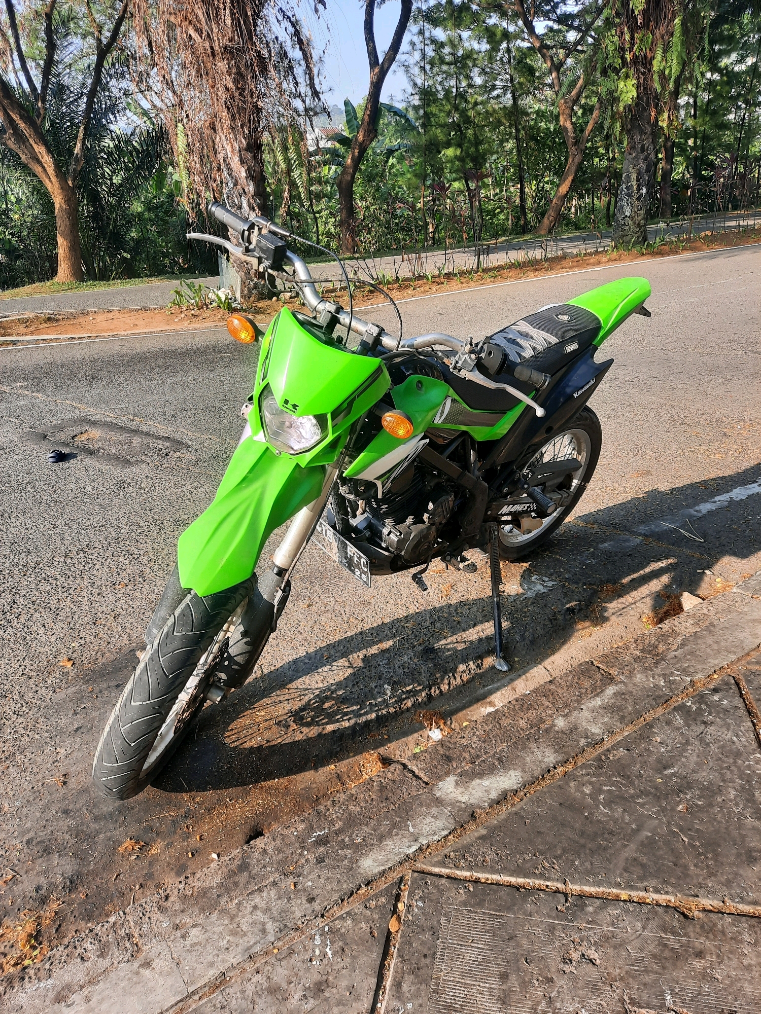 A bright green Kawasaki motorcycle is parked on the side of a paved road in Babakan Madang, Indonesia. The motorcycle is facing the viewer and angled slightly to the left. It has a black seat with a patterned cover and a black rear fender. The front of the motorcycle features a prominent green fender and a headlight. The bike is supported by a kickstand. The road surface is asphalt, with some debris and a white line visible. To the right of the road is a concrete curb and sidewalk, also with debris. Behind the motorcycle, a dense line of trees and foliage forms the natural environment. The sky above is a clear light blue. The lighting suggests it is daytime, with shadows cast by the motorcycle and surrounding trees.