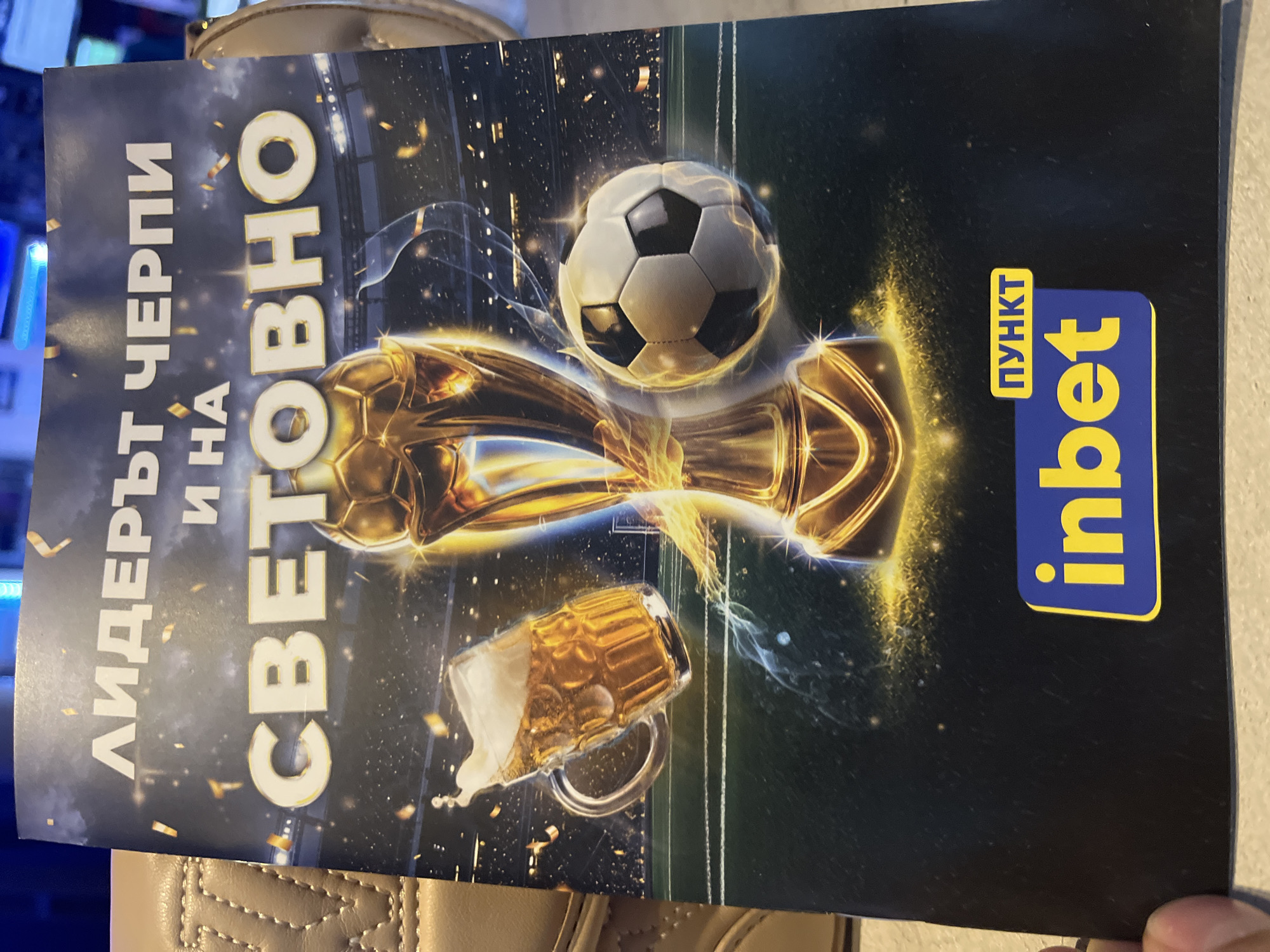 The image shows a close-up of a flyer or poster with a dark background and a soccer theme. At the top, there are white Cyrillic letters that appear to say "ЛИДЕРЪТ ЧЕРПИ И НА СВЕТОВНО". Below this text, a golden soccer trophy is prominently displayed in the center. To the left of the trophy is a glass mug filled with beer, with foam spilling over the top. To the right of the trophy is a soccer ball. The background depicts a stadium filled with spectators, illuminated by lights. At the bottom of the poster, there is a blue and yellow logo that reads "inbet" with smaller yellow text above it saying "ПУНКТ". The flyer is partially visible, with the edge of a light-colored textured material on the left side of the frame. The overall impression is promotional, likely for a sports-related event or establishment. The location is Unknown, Unknown.