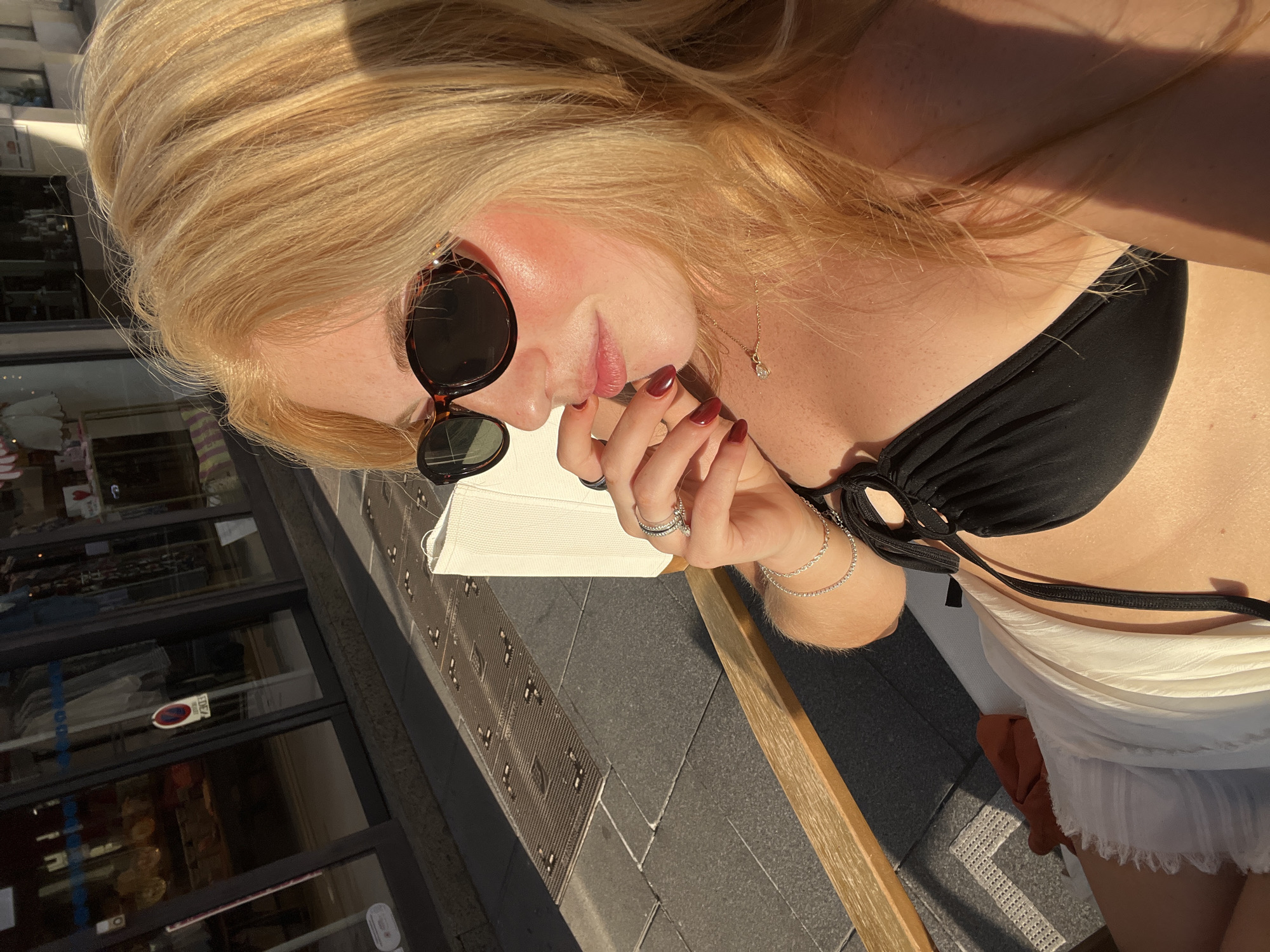 A sunlit, rotated overhead shot depicts a woman in Cannes, France, wearing a black bikini top and white skirt, accessorized with sunglasses, rings, a bracelet, and a necklace. She is sitting at a table with a white cushion, her hand near her face. In the background is a street scene, with a glass-fronted building reflecting the outside environment. The area appears to be paved, with linear architectural features. The scene is captured during daylight hours.