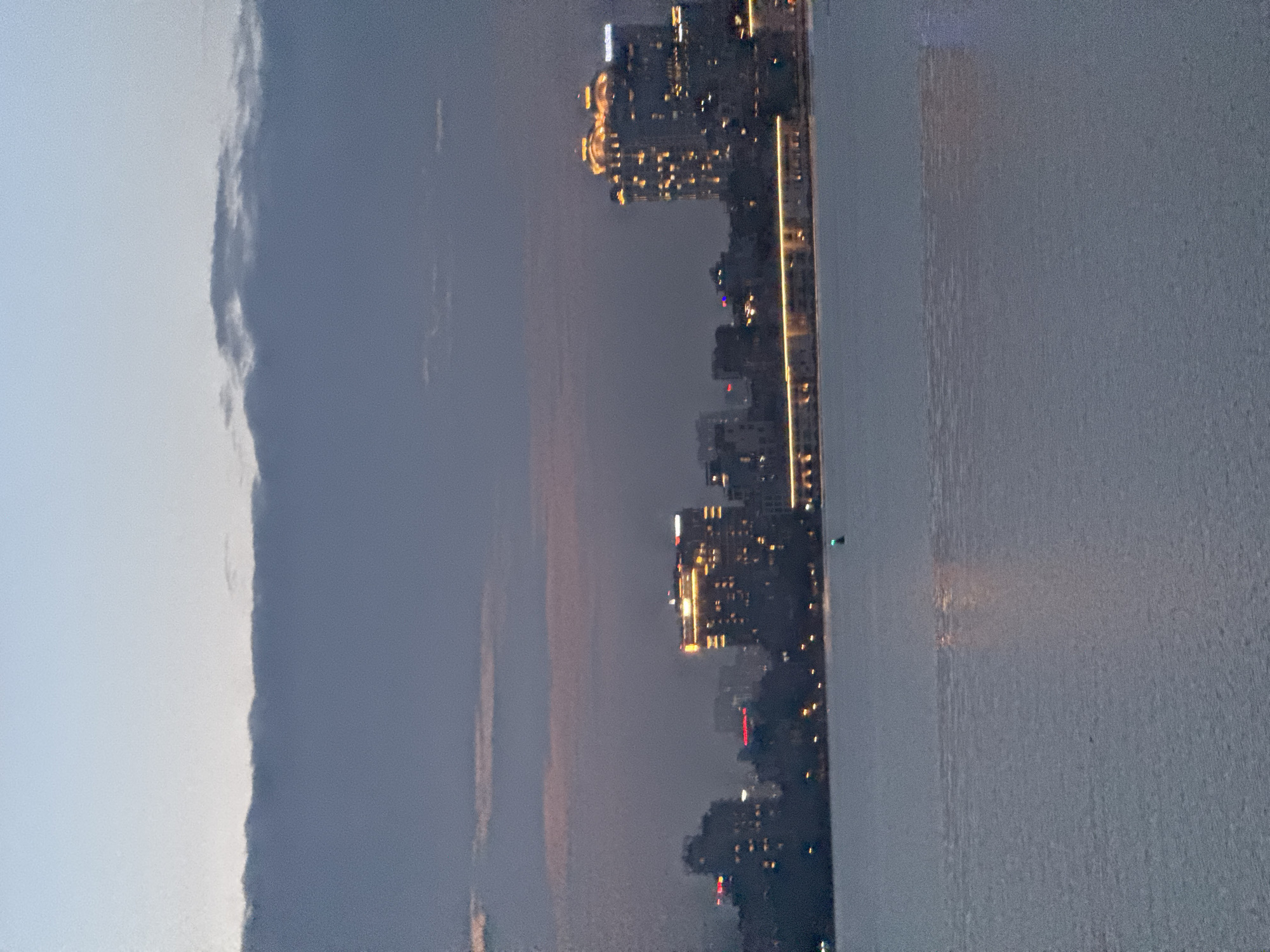 This image depicts a serene evening scene in Hanoi, Vietnam, likely near West Lake (Hồ Tây). The sky is a dusky blue, transitioning from a lighter shade at the top to a darker hue as it approaches the horizon. A band of soft, hazy clouds stretches across the upper portion of the image, tinged with faint hints of pink and orange, suggesting the lingering light of sunset or the approach of twilight.
Below the sky, the cityscape of Hanoi emerges from the gloom. Numerous buildings, ranging from mid-rise structures to taller, more prominent towers, are illuminated with warm, golden lights, indicating that it is nighttime or late evening. The lights create a soft glow against the darkening sky and reflect subtly on the water. Among the illuminated buildings, one particularly tall, ornate tower stands out with its crown-like upper floors brightly lit. Another building on the left is also well-lit, showing distinct window patterns.
The foreground is dominated by a large body of water, likely West Lake, which appears calm and still. The water's surface is a muted gray, reflecting the ambient light from the city and the sky. A faint shimmer of light is visible on the water's surface, possibly from the city lights or a distant buoy. A single green navigational buoy can be seen on the water, marking a point in the lake. Trees and silhouetted foliage are visible along the shoreline, adding a touch of natural landscape to the urban vista. The overall impression is one of tranquility and urban beauty as night falls over Hanoi.