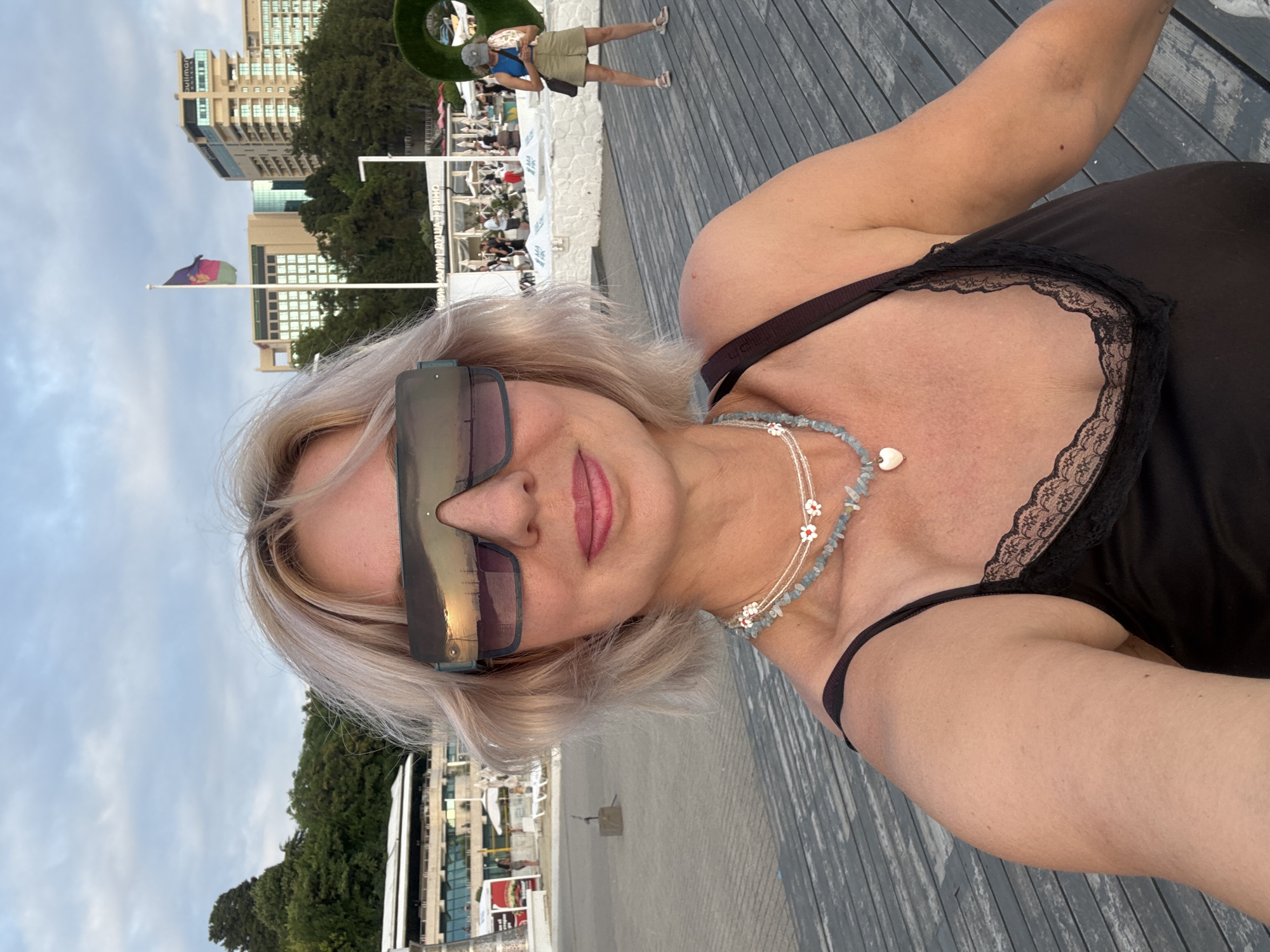 A woman with blonde hair and large sunglasses is taking a selfie on a wooden promenade. She is wearing a black camisole with lace trim and two necklaces, one with blue beads and a heart-shaped pendant, the other with white flower charms. In the background, there is a beach, a building with awnings, several trees, and a flagpole with a flag. Beyond that, there are tall buildings. The sky is overcast. The scene appears to be a waterfront area in Sochi, Russia, possibly near a beach resort.