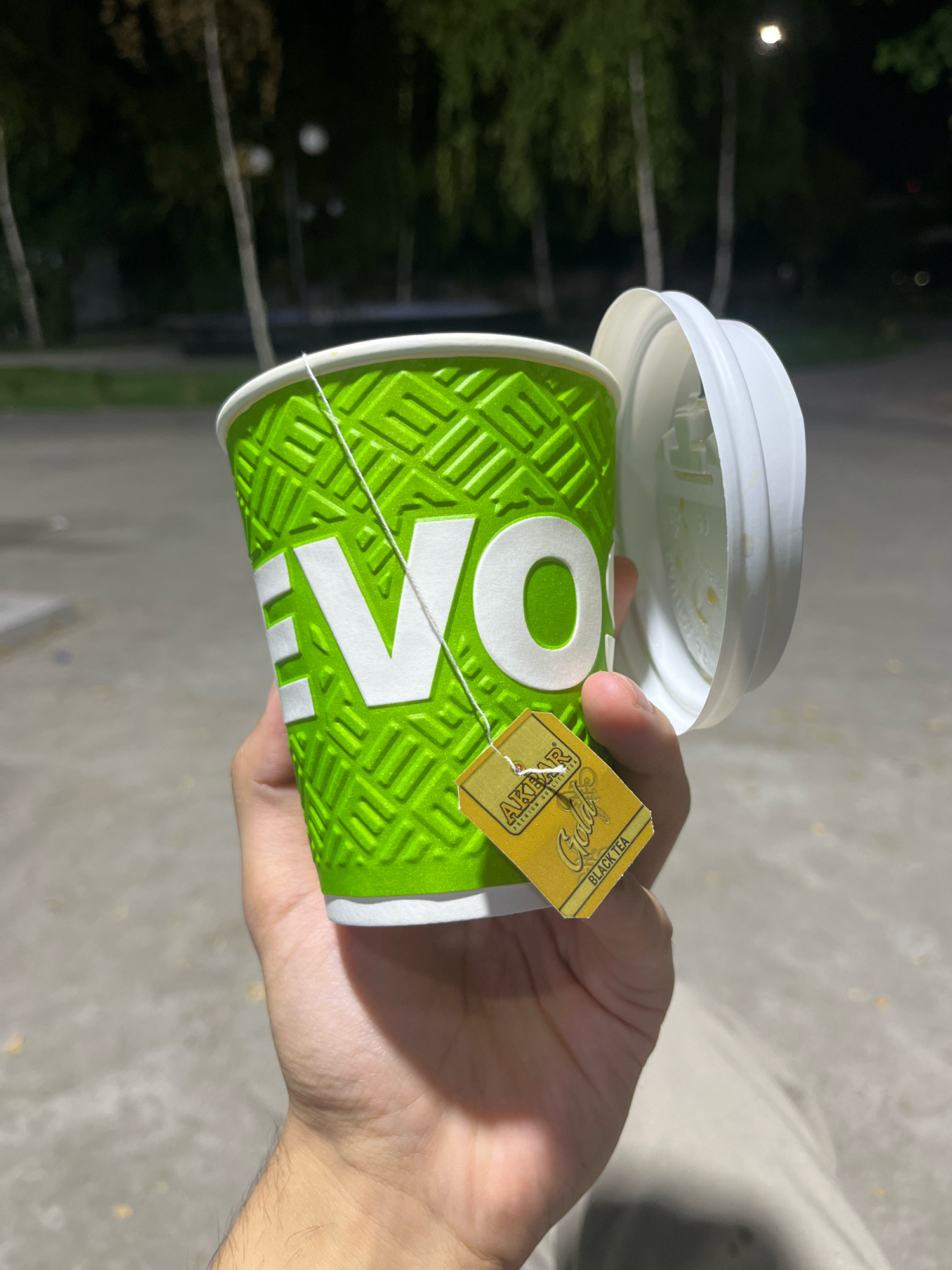 A person's hand holds a vibrant green paper cup with the embossed text "EVO". A white string from a tea bag is draped over the cup, with the yellow tea tag, featuring the "AKBAR Gold" logo and "BLACK TEA" text, resting on the side of the cup. A white plastic lid is partially open, revealing the inside of the cup. The background shows trees and streetlights, suggesting an outdoor setting at dusk or night, possibly in Tashkent, Uzbekistan. The ground appears to be paved.