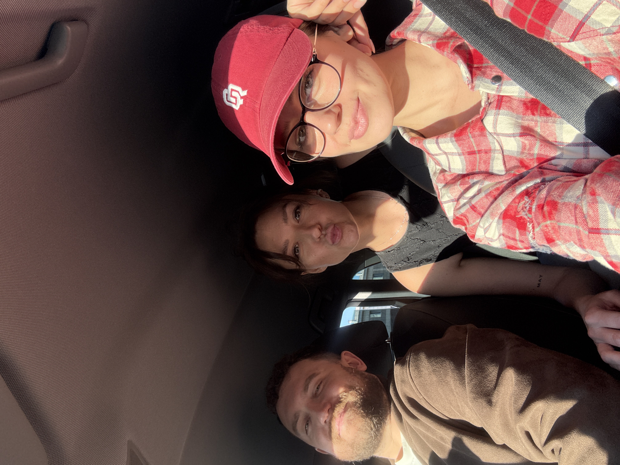 A selfie taken from the ceiling of a car shows three people. The person at the top, wearing a red baseball cap and glasses, has a seatbelt across their chest. Below them, a second person is making a kissing face. The third person, at the bottom, has a beard and is smiling. The location is unknown, and there is no evidence of being in the city of Krasnoyarsk, Russia.