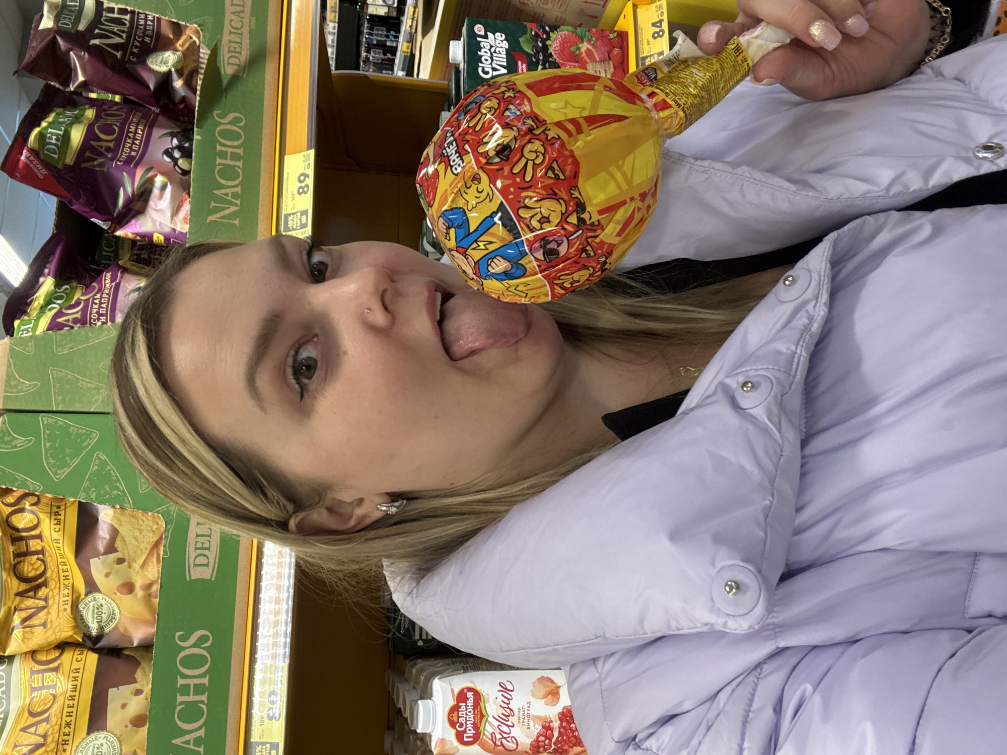 A woman is holding a large lollipop in front of her face with her tongue sticking out. She is wearing a light purple puffer jacket and has blonde hair. The background shows shelves stocked with groceries, including "Nachos" and juice boxes labeled "Сады Придонья" (Sady Pridonya). Prices on the shelves suggest a retail setting, likely a grocery store or supermarket. The location is likely Chaikovsky, Russia, based on the Cyrillic writing and the brand of juice boxes.