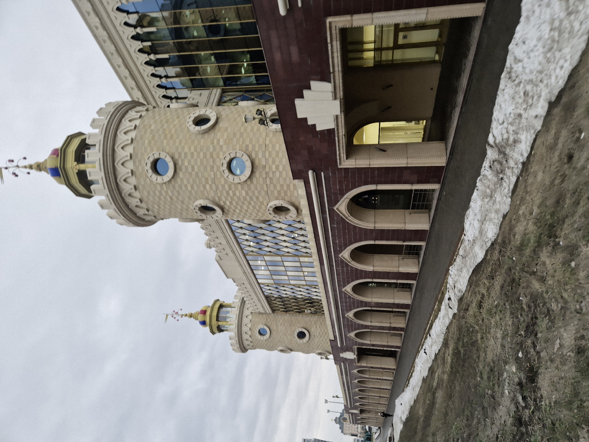 The image presents the architectural structure of the Kazan Puppet Theater located in Kazan, Russia. The building exhibits a whimsical and elaborate design, featuring castle-like turrets with circular windows, ornate decorative moldings, and multi-colored spires topped with weather vanes.
The building's exterior includes a combination of light-colored stone brickwork and dark red brick sections, marked by rounded arches and rectangular windows. The facade incorporates large glazed areas reflecting the sky and surrounding environment.
The theater is situated adjacent to a paved walkway, separated from the building by a narrow concrete strip. The walkway is bordered by patches of snow and grass, indicating a transition from winter to spring. The sky appears overcast.
The image is taken from a ground-level perspective, with the camera angled upwards to capture the height and detailing of the building. Distant urban structures and streetlights suggest a city environment. There is one person walking along the right side of the image.