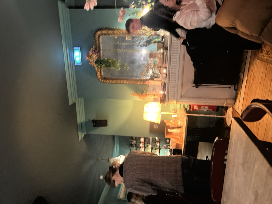 An interior scene showcases a green-toned room with a large ornate mirror above a white fireplace mantle. A glowing lamp illuminates the area. Two people are seated on a sofa, one wearing white and the other a dark outfit. In the foreground, a person wearing a grey patterned vest stands by a table with chairs. A bar area is visible in the background, stocked with various items. A green illuminated sign is on the wall near the mirror.