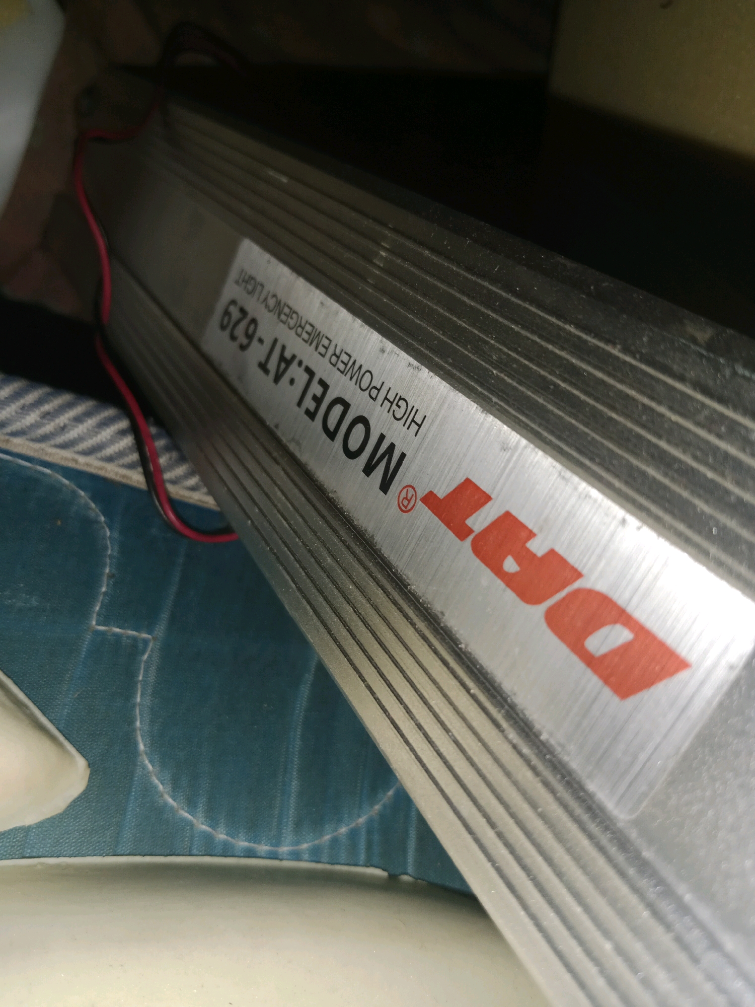 The image shows a close-up of an emergency light with the brand name "DAT" and model number "MODEL: AT-629 HIGH POWER EMERGENCY LIGHT" visible on a silver casing. The light is a rectangular, elongated object with a ribbed metallic exterior, and red and black wires are attached to it. The emergency light is resting on a blue and white striped surface that appears to be fabric, possibly part of a mattress or cushion. The background is dark and out of focus. There are no people visible in the image. The location is not specified in the image or its metadata.