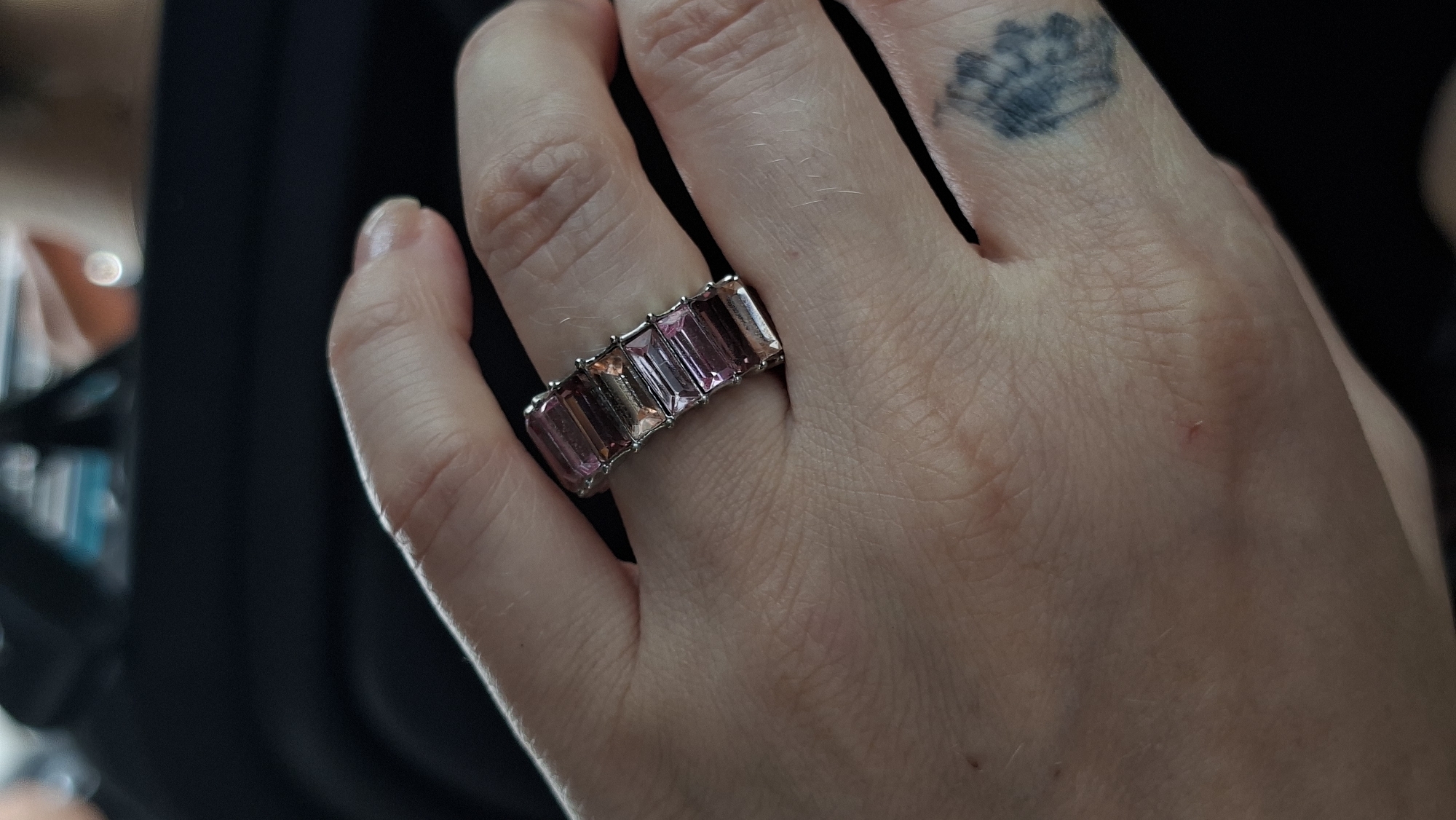 The image shows a close-up of a hand wearing a ring. The ring is made of silver and features five rectangular-cut gemstones in shades of pink and clear. The hand has fair skin with some visible veins and light stubble on the knuckles. There is a tattoo visible on the back of the hand, partially obscured by shadow. The background is dark and out of focus, suggesting an indoor setting, possibly a car interior given the dark material and slight curvature. There is no indication of the city of Myadzyel, Belarus in the image.