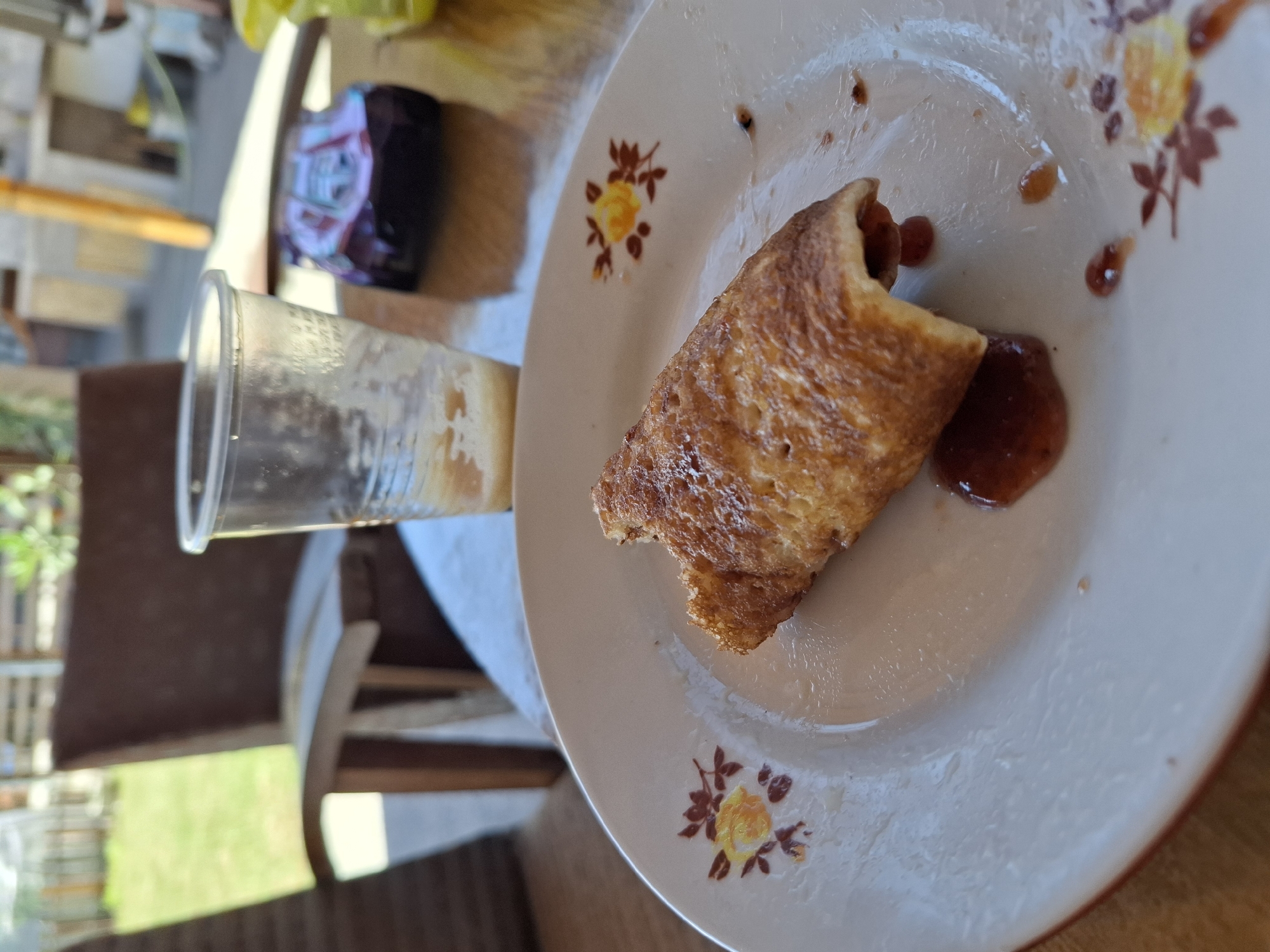 A close-up shot focuses on a single, partially eaten rolled pancake or crepe on a white plate with floral decorations. The food item has a golden-brown, slightly uneven surface and appears to be filled with a dark red jam or fruit filling that has spilled out onto the plate. Several small droplets of the same jam are scattered around the food. The plate is positioned on a wooden table. In the blurred background, a clear plastic cup with remnants of a beverage is visible, along with a toy car. Further back, a wooden chair and a grassy area suggest an outdoor or semi-outdoor setting, possibly a backyard or patio. The overall scene implies a casual meal or snack, with the toy car and outdoor elements hinting at a family-friendly environment. The image does not contain any identifiable landmarks specific to Galabovo, Bulgaria, beyond the general context of a domestic setting.