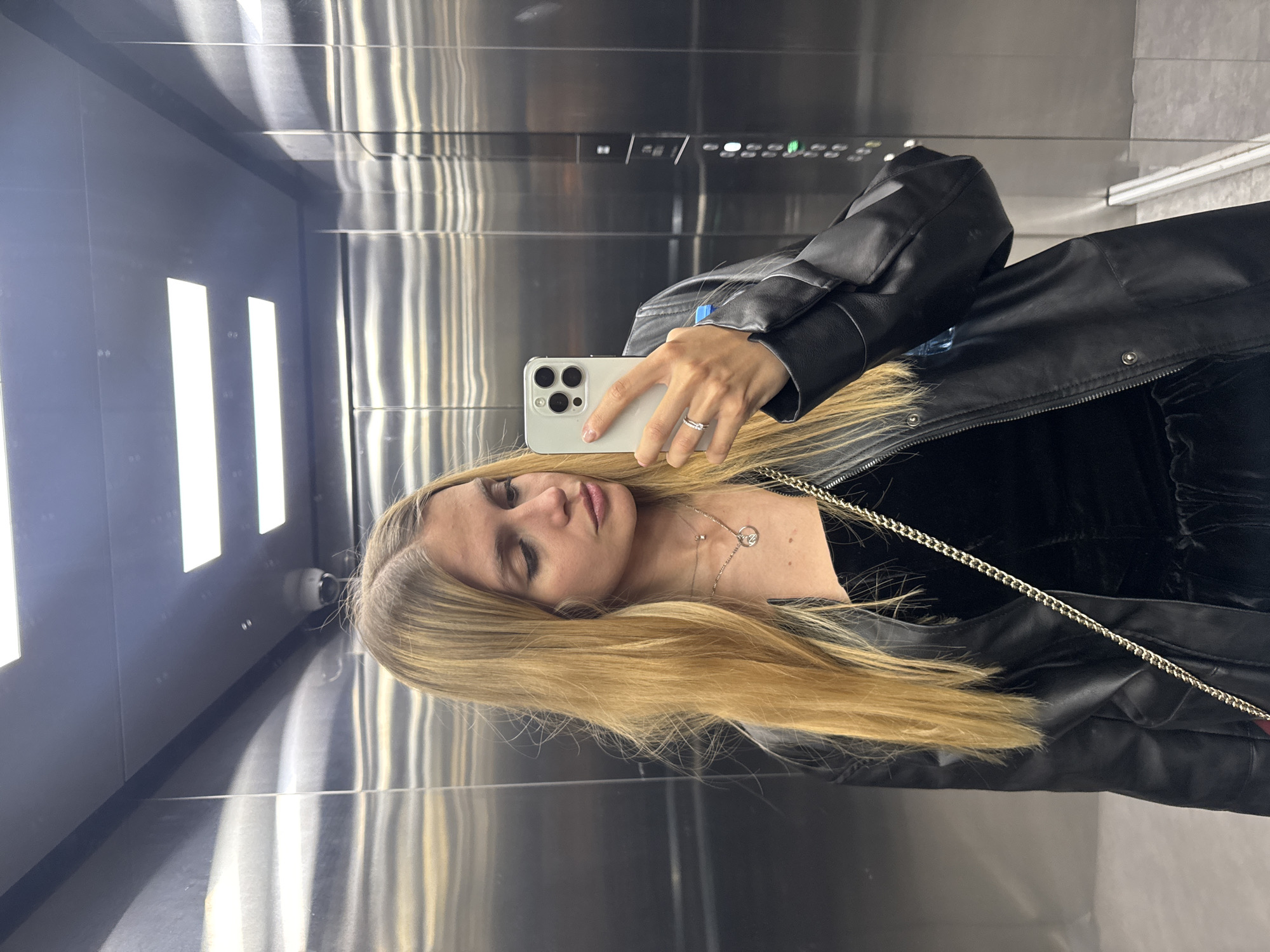 A woman is taking a selfie in an elevator in an unknown city. She has long blonde hair and is wearing a black leather jacket over a black top, with a gold chain necklace and a ring on her finger. She is holding an iPhone in her hand. The elevator interior is stainless steel. The elevator panel with floor numbers is visible above her. There are lighting fixtures running vertically along the wall. A security camera is mounted on the wall.