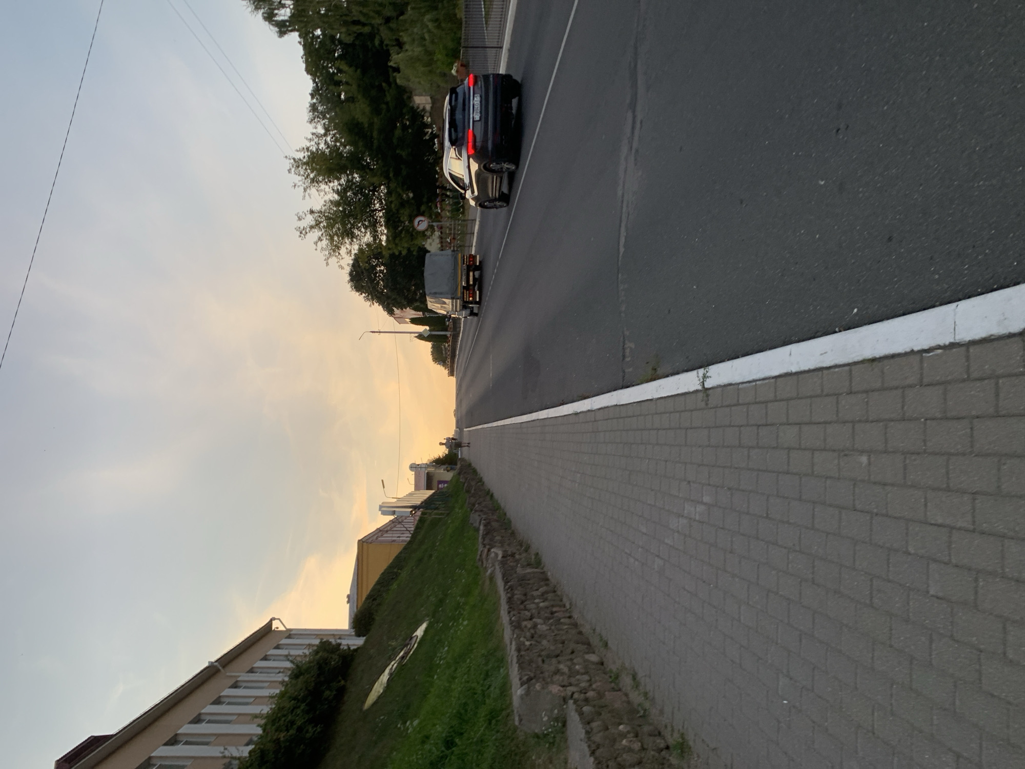An exterior, eye-level, vertical, full shot shows a paved sidewalk and an asphalt road in the city of Kapyĺ, Belarus. The scene captures the golden hour light of dusk, with a soft glow illuminating the sky.
On the left side of the image, a light-colored building with multiple windows stands on a grassy slope. The slope is bordered by a low, stone retaining wall. The sidewalk, made of light gray interlocking pavers, runs along the base of the slope. A single person, appearing as a silhouette, is walking on the sidewalk in the distance.
The asphalt road occupies the right side of the image, curving slightly uphill. A dark-colored SUV with its taillights illuminated is driving on the road, followed by a truck with a grey tarp covering its cargo. Trees with dense green foliage line the right side of the road, with a dark metal fence visible behind them. Overhead power lines are also visible against the pale sky. The overall impression is of a quiet urban street during the evening transition.