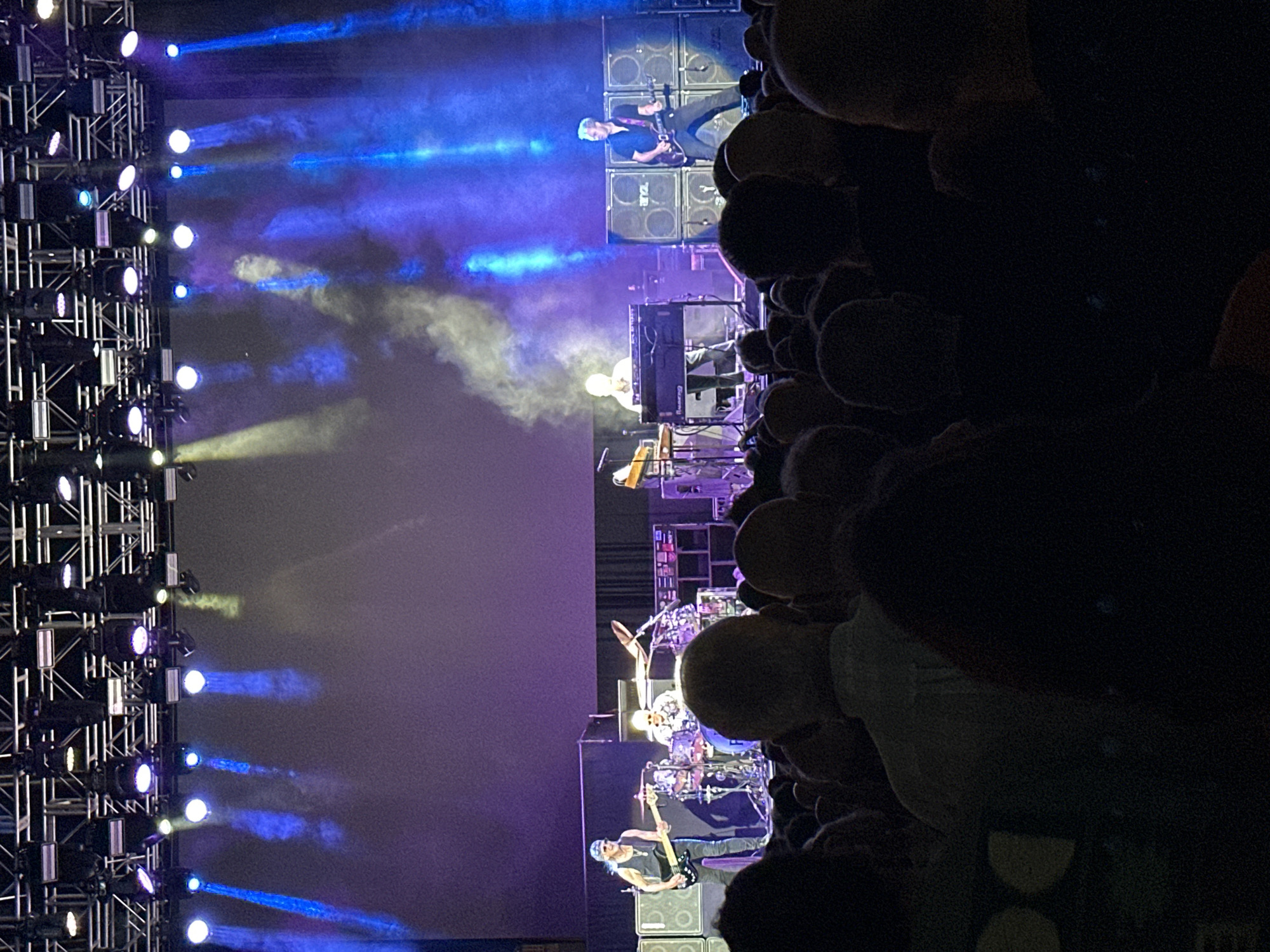 The image shows a live music concert taking place on a stage. A band is performing, with members playing various instruments including guitar, drums, and keyboards. The stage is illuminated by dramatic lighting, with blue and purple spotlights creating beams and haze effects. In front of the stage, a large audience is visible, their faces mostly obscured by darkness and the foreground. The overall atmosphere suggests an energetic performance. The image does not provide any specific details that would allow for the identification of the city of Jerez, Spain, or any other specific location.