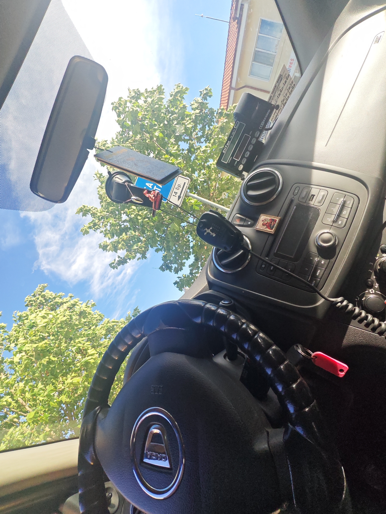 The image shows the interior of a Dacia car, likely parked in Nova Zagora, Bulgaria. The steering wheel with the Dacia logo is prominent, along with the car's dashboard. A radar detector and a phone mount are attached to the windshield. A small religious icon is affixed to the dashboard. Outside the windshield, trees and a building are visible, with a parking sign reading "14 бр" (14 pieces). A taximeter "EllTax TA-100/F1" is seen mounted to the dashboard. The label "ГРОВА ТАКСИ ЕООД" (GROVA TAXI Ltd.) is visible, along with taxi fare information. A red key tag is attached to something near the steering wheel.
