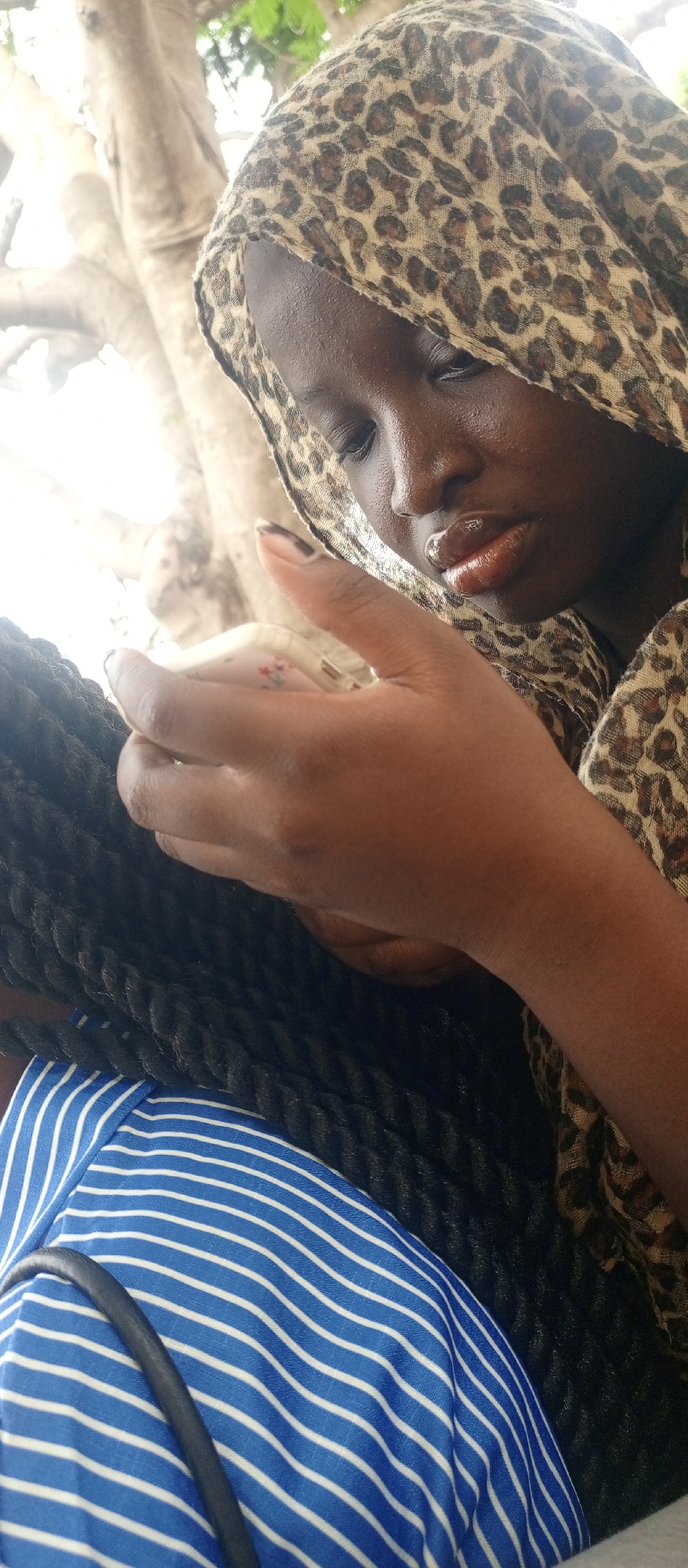 A close-up, high-angle shot captures a person, likely female, in Keffi, Nigeria, during the daytime. The individual has dark skin and is wearing a light-colored head covering with a brown and black leopard print pattern, which partially obscures their face. Their eyes are cast downwards, focusing on a light-colored smartphone held in their hands. The phone appears to have a pale pink or white case with a subtle floral design. The person's lips are prominent and appear glossy, possibly from lip gloss. Their fingernails are painted with a dark polish.
Below their hands and the leopard print fabric, a mass of thick, black braided hair is visible, suggesting a hairstyle of twists or braids. The person is also wearing a garment with a distinct blue and white horizontal stripe pattern. A dark strap, possibly from a bag, rests over the striped fabric.
The background is brightly lit and blurred, showing what appears to be light-colored tree trunks and some green foliage, indicating an outdoor or semi-outdoor setting. The strong natural light suggests it is sunny. The overall mood is one of focused engagement with the smartphone.