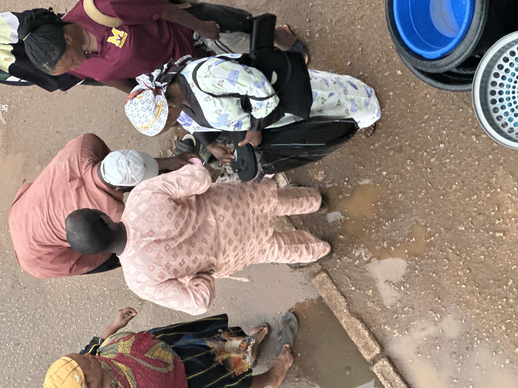 An aerial view shows a group of people gathered on a muddy, uneven surface in Bukuru, Nigeria, likely a street or market area. Several individuals are dressed in traditional Nigerian attire, including a person in a pink kaftan and another in a patterned white and blue outfit. One person wears a maroon shirt with an "M MADISON" logo. The ground is wet and appears to have standing water in puddles, indicating recent rain or poor drainage. Objects visible include black plastic buckets, one containing a blue basin, and a sieve-like metal object. The interaction seems casual, with people engaged in conversation or tending to items.