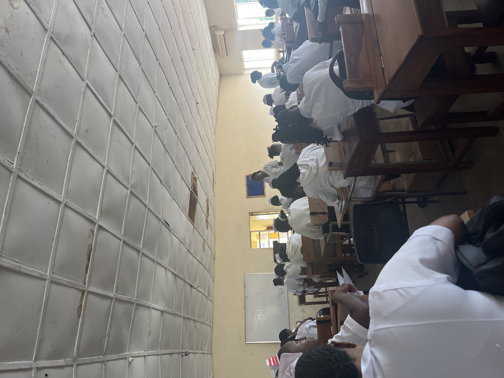 The image depicts a classroom setting in Jos, Nigeria, likely within an educational institution. The room is filled with students, predominantly young women, dressed in white uniforms, suggesting they are nursing students or engaged in a medical-related program. They are seated at wooden desks, facing the front of the classroom.
In the front of the room, a whiteboard is visible with some writing on it, though it is partially obscured. To the right, windows allow natural light to enter, and an air conditioning unit is mounted high on the wall. The ceiling appears to be a drop ceiling with visible water stains in some sections, indicating potential disrepair.
The students are in various states of attention, with some appearing to listen to an instructor (partially visible at the front), while others are looking down at papers or their desks. The overall atmosphere suggests a typical academic lecture or study session. The presence of the whiteboard and desks clearly defines the space as a place of learning.