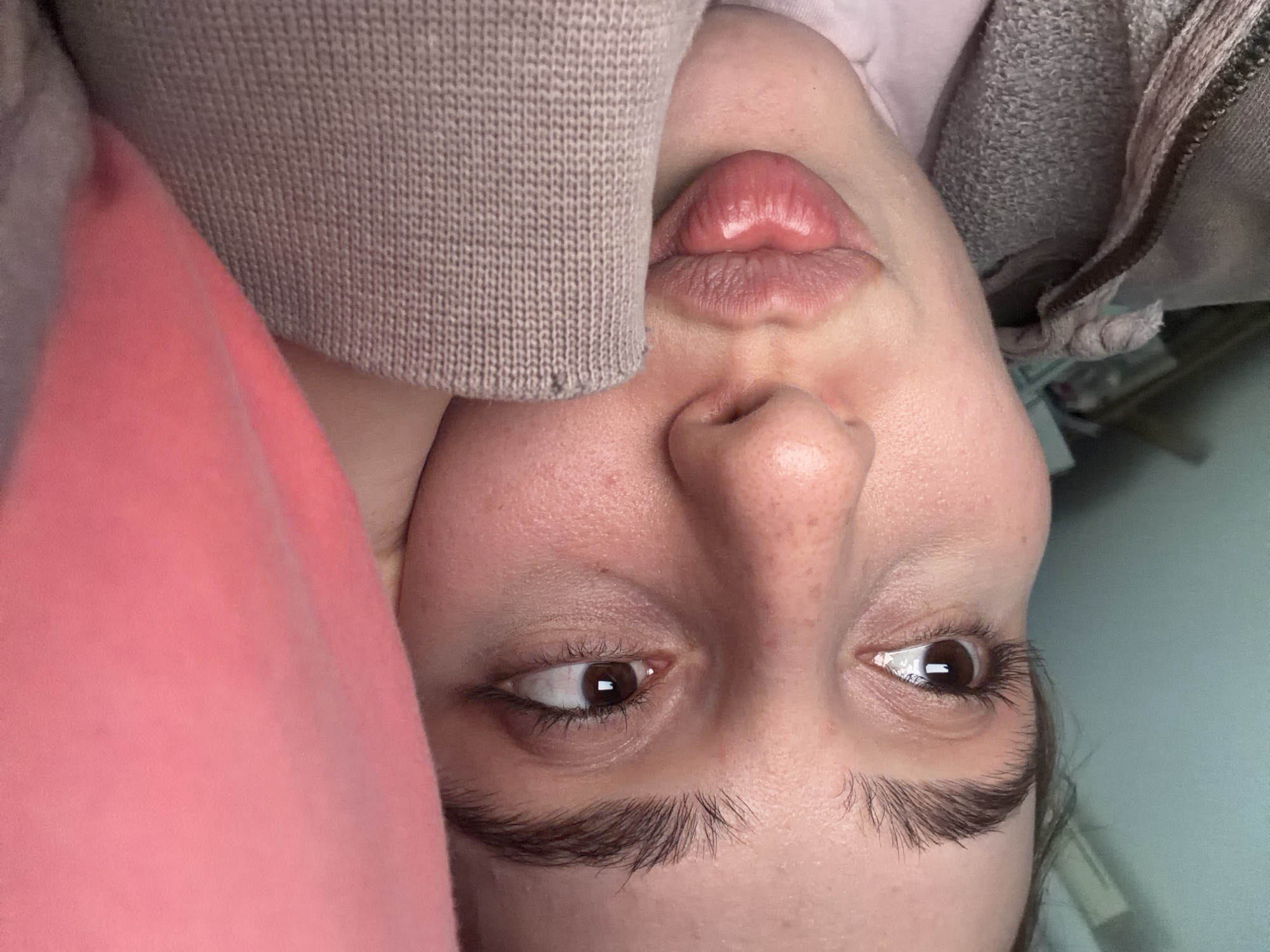 The image presents a close-up, upside-down portrait of a person's face, captured in an indoor setting in Strasbourg, France. The subject, likely female, is positioned centrally with their head inverted, suggesting a relaxed or playful pose.
The person's facial features are prominent. Their lips are full and glossy, puckered into a distinct pout. Their nose is visible in the lower center of the inverted face. Both dark brown eyes are open and looking towards the viewer's left (which is the subject's right). Long, dark eyelashes frame the eyes, and their eyebrows are thick and natural. The skin has a visible texture, showing pores and minor imperfections. A hint of dark hair is visible near the person's right eye.
Various fabrics surround the individual. A light beige or pale brown knitted material, possibly a sweater sleeve or blanket, covers a portion of the left side of the inverted face. To the right, a soft, vibrant pink fabric, potentially a blanket or clothing item, partially obscures the lower left of the frame. In the upper right corner, a dark grey, textured fabric, resembling a hoodie or sweatshirt, can be seen. The background behind the person's right eye shows an out-of-focus light blue surface, consistent with an indoor wall.
The image conveys a casual and possibly playful mood, characteristic of a personal snapshot or selfie. There is no visible text in the media file.