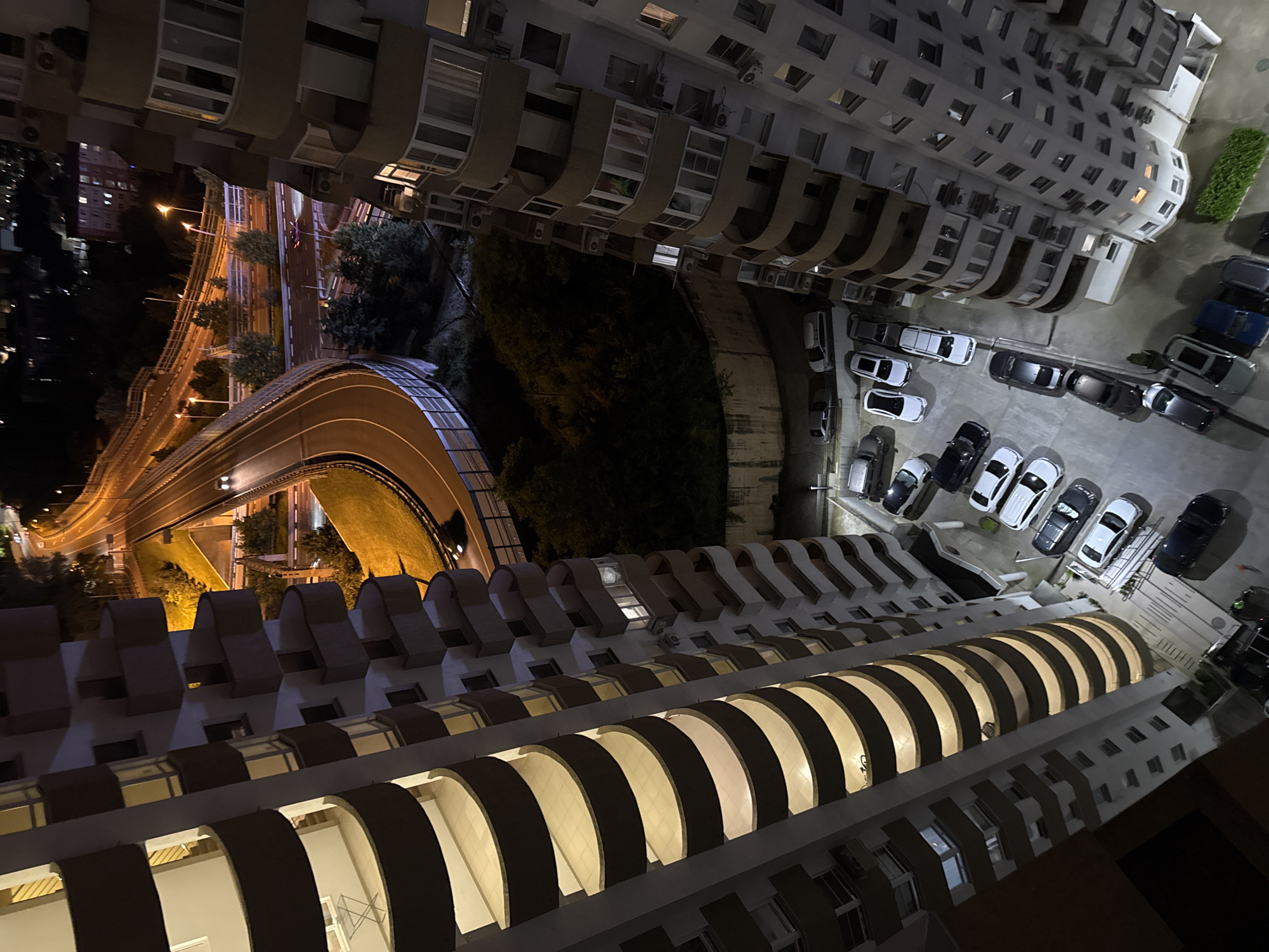 This high-angle, downward-looking image captures a nighttime urban scene in Sochi, Russia. Two tall, multi-story residential buildings frame the composition. The building on the left, running vertically along the left edge, features numerous curved, light-colored balconies with dark undersides, each illuminated from within, creating a distinct striped pattern ascending the facade. A small white outdoor table is visible on one of these balconies. The building on the right, also multi-storied, exhibits angular, recessed balconies, many with windows and some featuring external air conditioning units.
Between these two structures, a complex multi-level highway interchange is visible, brightly illuminated by numerous streetlights, casting an orange glow on the asphalt and surrounding grassy embankments. Several vehicles with active headlights and taillights traverse the curved roadways, indicating movement. Below the elevated highway structures and to the left of the right building, a dark, densely vegetated area is present, bordered by a concrete retaining wall at its base.
In the foreground and center-bottom, a large outdoor parking lot, illuminated by ambient light, contains numerous parked passenger vehicles of various models and colors, arranged in rows. No people are discernible in the image.