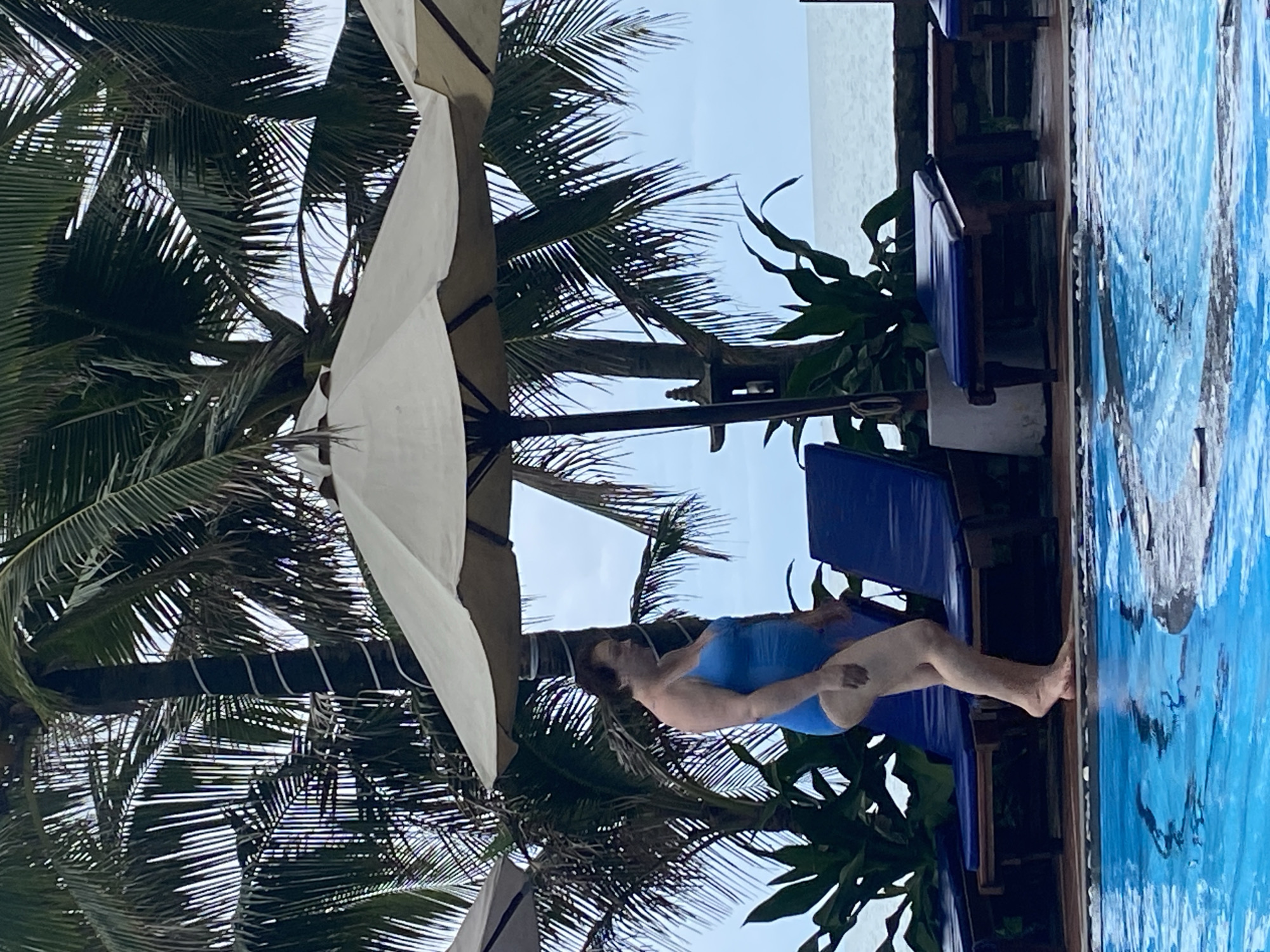 A woman in a blue swimsuit is lying on a blue lounge chair by a pool, with palm trees and a white umbrella in the background. The location is in or near Phu Thuy Ward, Vietnam. The pool appears to be outdoor. The woman is facing away from the viewer, and her feet are near the edge of the pool.