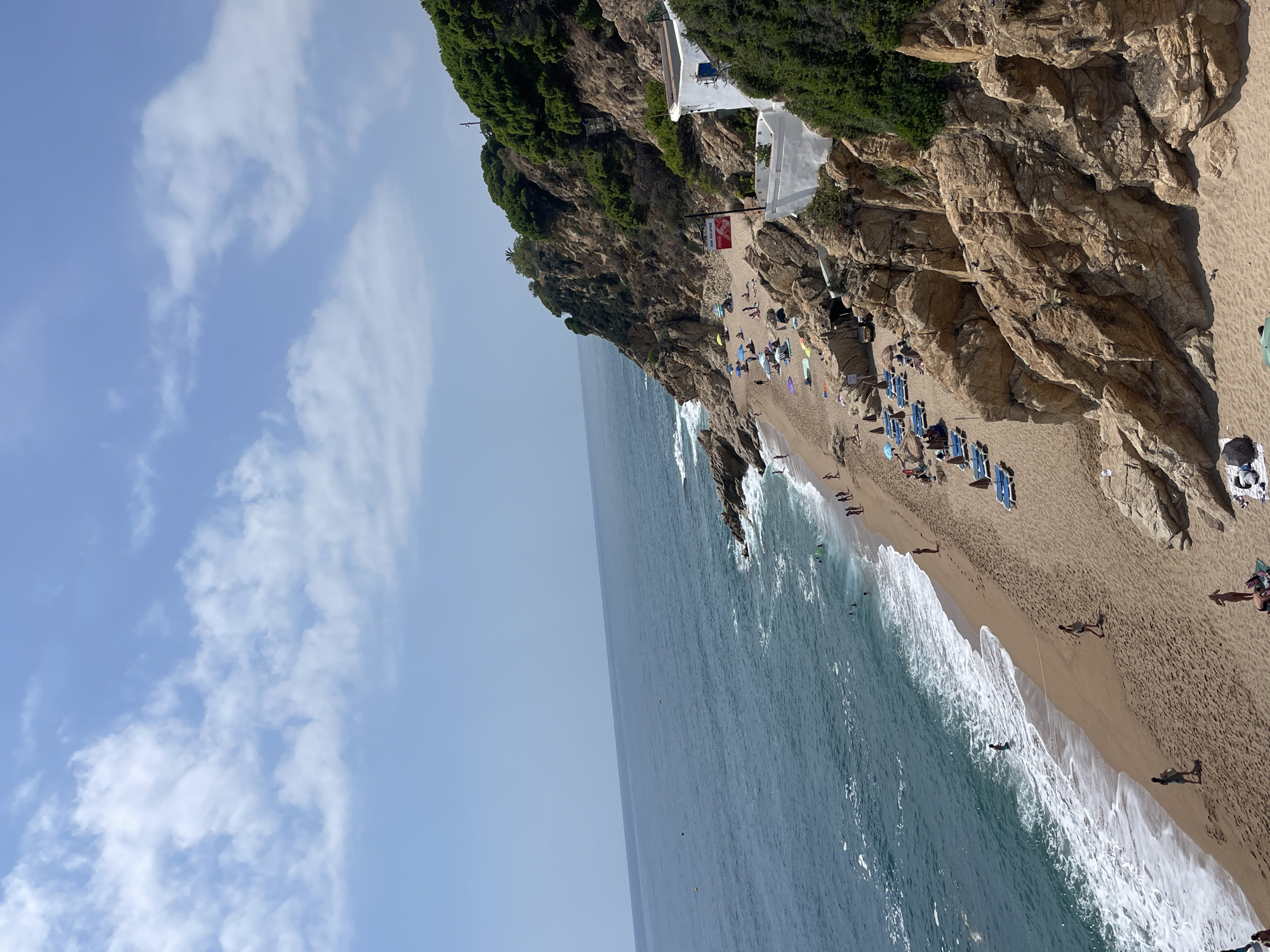 The image captures a scenic view of a beach in Calella, Spain, with the Mediterranean Sea on the left and a rocky, tree-covered cliff on the right. The beach is populated with sunbathers relaxing on lounge chairs under umbrellas, and individuals swimming or walking along the shore. The water is a clear blue-green, with gentle waves breaking on the sandy beach. The cliff face is rugged and natural, with dense green foliage. A white building with blue accents is visible near the top of the cliff, and a red sign that says "BAR ROCAPRI" with a Coca-Cola logo is attached to a pole on the beach. The sky is partly cloudy, with fluffy white clouds scattered across a bright blue expanse. The overall scene conveys a sense of a peaceful and lively beach day.