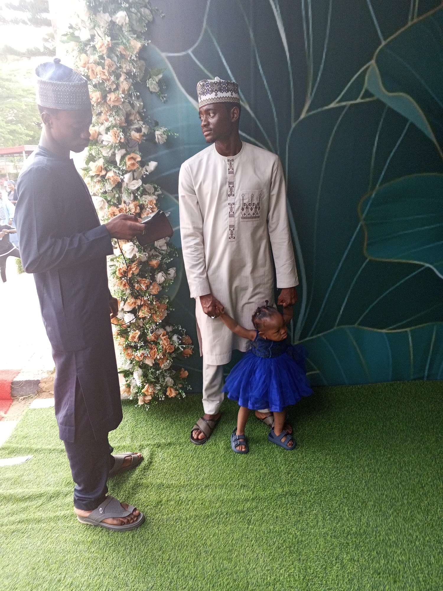 The image depicts an outdoor scene, likely in the city of Abuja, Nigeria, featuring three individuals. In the foreground left, a man wearing a dark blue traditional outfit and a patterned cap is holding a small black object, possibly a phone or wallet, in his left hand. He is looking down at it. Beside him and slightly behind, another man in a light-colored traditional outfit and a patterned cap stands, holding the hand of a young child. The child, a toddler, is dressed in a bright blue puffy dress and is looking towards the ground. The background features a floral arrangement with white and orange flowers and a large teal-colored backdrop with a leaf-like pattern. The ground is covered in artificial green turf. The lighting suggests it is daytime.