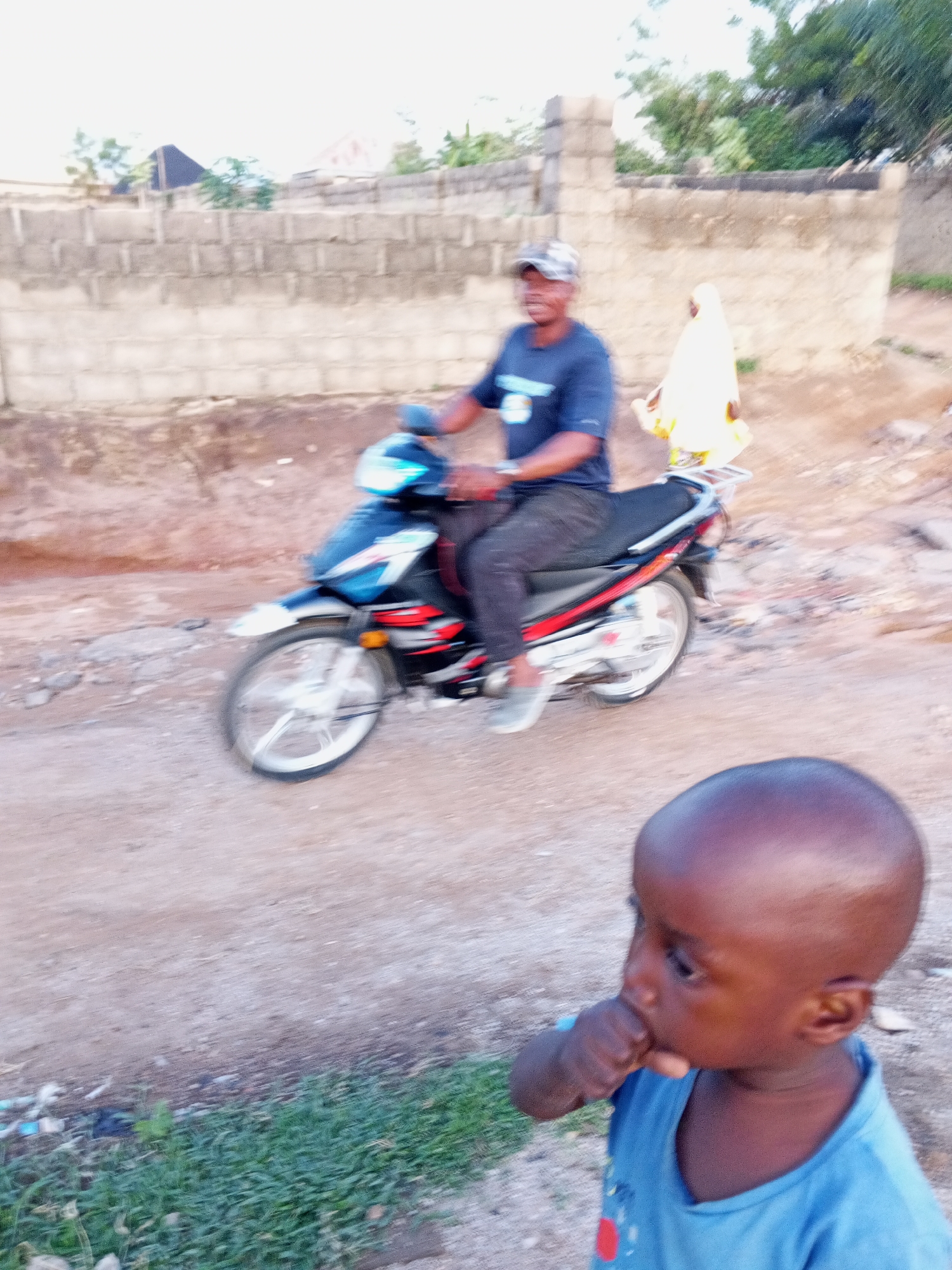 The image shows a man riding a black and red motorcycle on a dirt road in Zango-ndaji, Nigeria. The man is wearing a dark blue shirt, dark pants, a cap, and gray shoes. He has a passenger behind him with a white bag or cloth draped over them. In the foreground, a young child with dark skin is looking to the right, with their fingers near their mouth. The child is wearing a blue shirt. A low concrete block wall is visible in the background, along with some trees and buildings. The ground is dirt and grass.