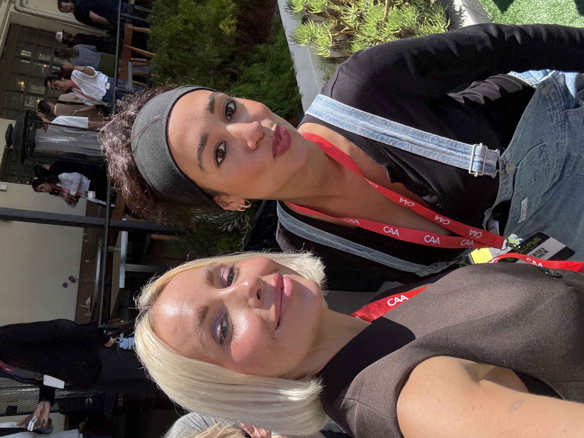 Two women are posing for a selfie outdoors. The woman at the top of the frame has dark hair, a black headband, and is wearing blue denim overalls over a black top. She is wearing a red lanyard with "CAA" printed on it. The woman at the bottom of the frame has blonde hair and is wearing a brown top. She is also wearing a red lanyard with "CAA" printed on it.
In the background, other people can be seen near tables and building structures. Green foliage is also visible.
The CAA lanyards suggest the location may be related to Creative Artists Agency. The presence of other people at tables implies a dining or social setting. The setting is likely in Los Angeles, United States of America, though specific landmarks are not identifiable.