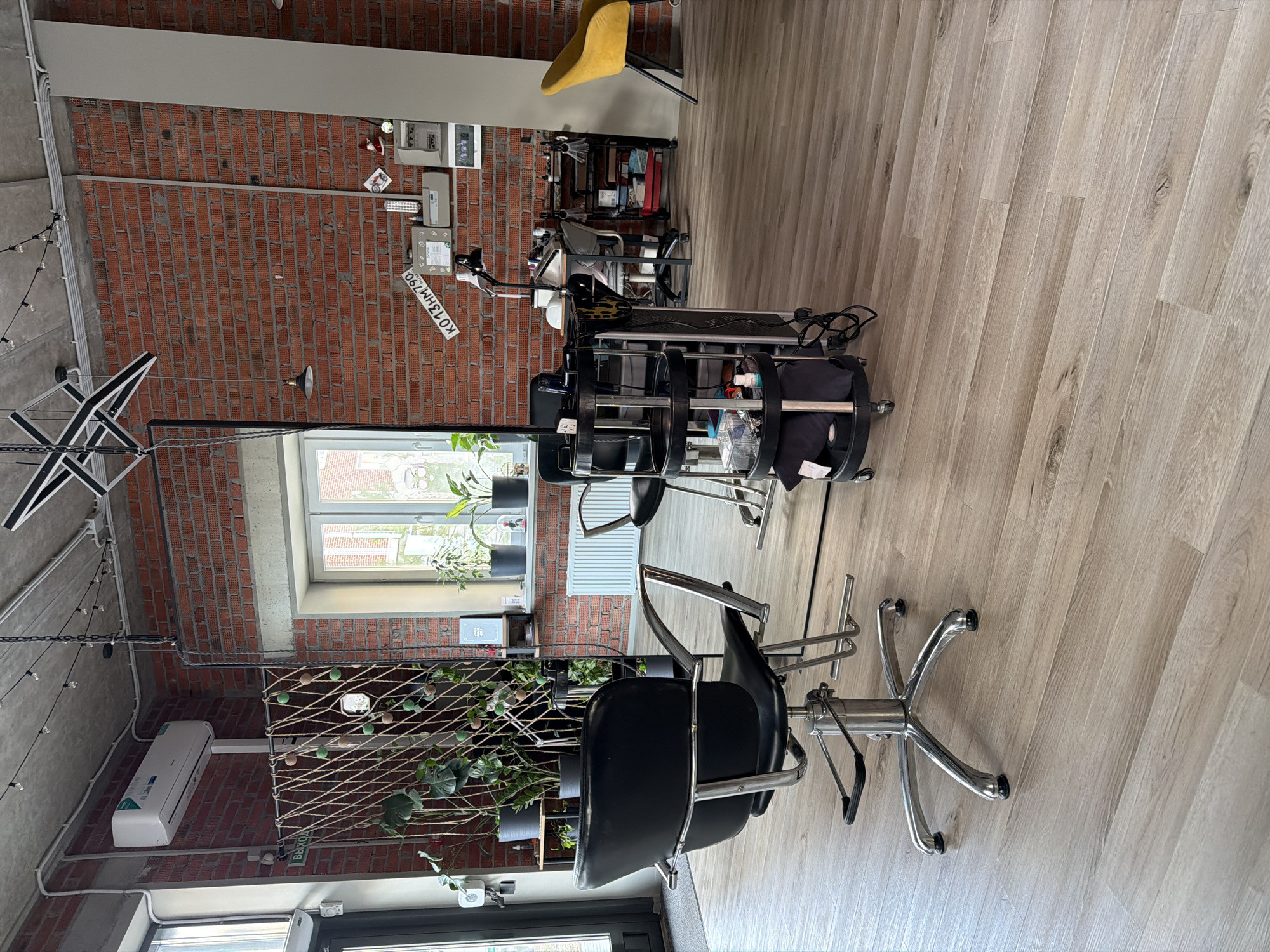 The image shows the interior of what appears to be a hair salon in Pervomaysky, Russia, based on the license plate visible on the wall ("K013HM790"). The salon has a modern, industrial style with exposed brick walls and wooden floors. A black salon chair is in the foreground, facing a mirror. A styling station is behind the chair, with a black cart holding various hair styling tools and products. A second, yellow chair is visible in the background. Potted plants are placed near a window and on a shelf with a rope net. An air conditioning unit is visible on the wall. Overall, the salon appears to be well-lit and organized.