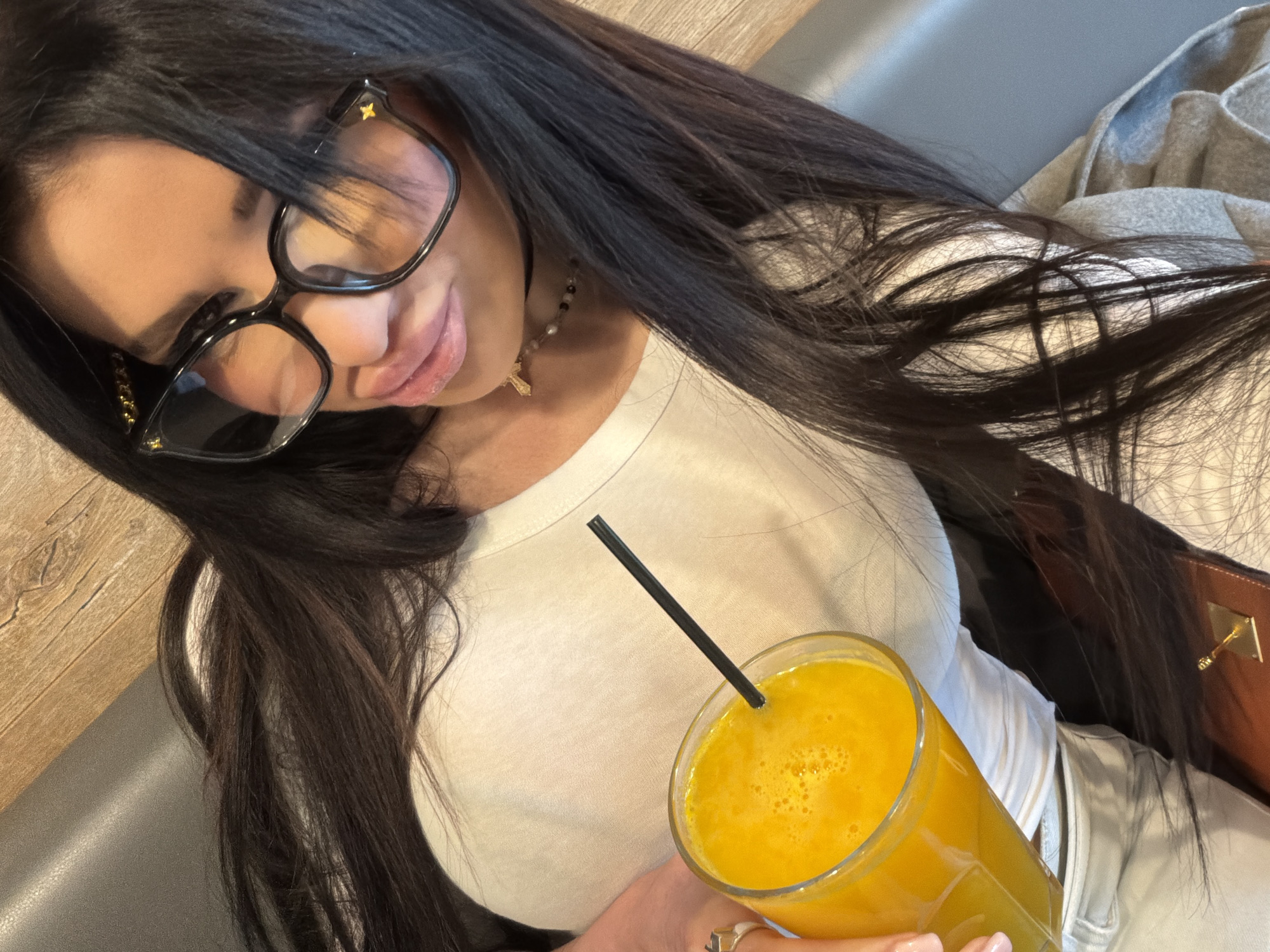 An indoor, tilted, close-up shot features a woman holding a glass of orange juice. The woman has long, dark, straight hair and fair skin. She wears black-framed glasses with small gold embellishments near the temples. Her lips are slightly parted in a soft, subtle smile, and she appears to be looking subtly towards the viewer. Around her neck, she wears a choker-style necklace with small beads and a prominent golden cross pendant.
She is dressed in a light-colored, possibly white or off-white, top. Her right hand, adorned with a ring featuring a light-colored stone, holds a clear glass filled with a vibrant orange beverage, likely juice, topped with some foam and containing a black straw.
The background shows elements suggesting an indoor setting: a wooden surface (possibly a table or counter) to the left, and gray upholstered seating. A gray blanket or jacket is partially visible behind her, and a brown bag or jacket is seen on the right. The image captures a casual and relaxed moment, possibly a selfie. The provided context indicates the location is Moscow, Russia, though this is not visually evident in the image itself.