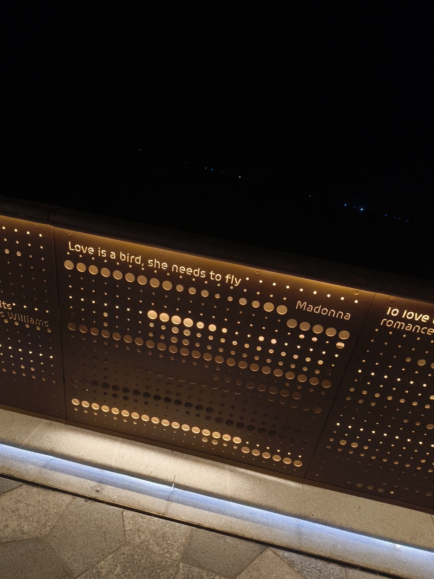 The image shows a perforated metal wall at night, illuminated from behind by lights that shine through the holes. The wall has text and circular patterns. The visible text reads "Love is a bird, she needs to fly", "Madonna", and "To love romance". There are also fragments of other words, like "hits" and "Williams". The wall is angled and appears to be part of a public space, possibly an outdoor walkway or seating area, given the stonework visible in the foreground. The sky is dark, with some distant lights visible. The setting is likely an urban environment. While specific landmarks in Phú Quốc, Vietnam are not identifiable from this image alone, such decorative illuminated walls are often found in entertainment districts, promenades, or at event venues in tourist areas.