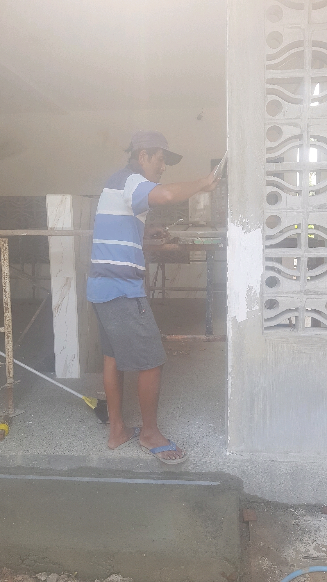 A man is plastering a wall in Sukodermo, Indonesia. He is wearing a blue and white striped t-shirt, grey shorts, and a baseball cap. The man is holding a trowel in his right hand and is applying plaster to a white decorative wall. The wall has a pattern of circles and waves. The man is standing on a concrete floor, and there is a pile of construction materials behind him. The sun is shining brightly, and the scene is somewhat hazy.