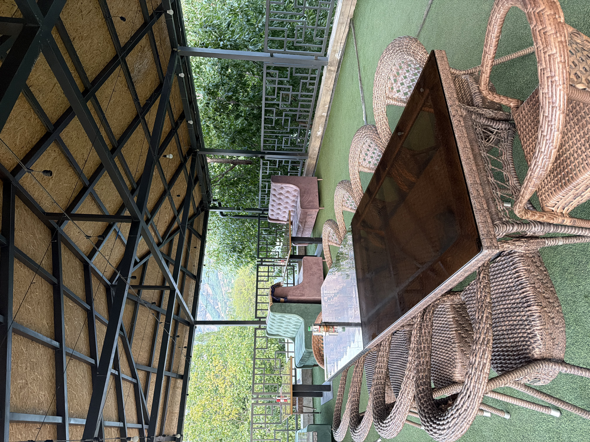This image shows an outdoor seating area of a restaurant or cafe, possibly located in Nanay, Uzbekistan. The area is covered by a roof supported by a dark metal frame and has an artificial turf floor. In the foreground, a rectangular dark glass-topped table is surrounded by several woven rattan chairs. In the midground, there are two tufted velvet sofas, one light green and one mauve, with a small table in between them. Further back, another similar sofa and a table are visible. A decorative metal railing runs along the edge of the seating area, overlooking a lush green landscape with trees and hills in the distance. There are no people present in the image.