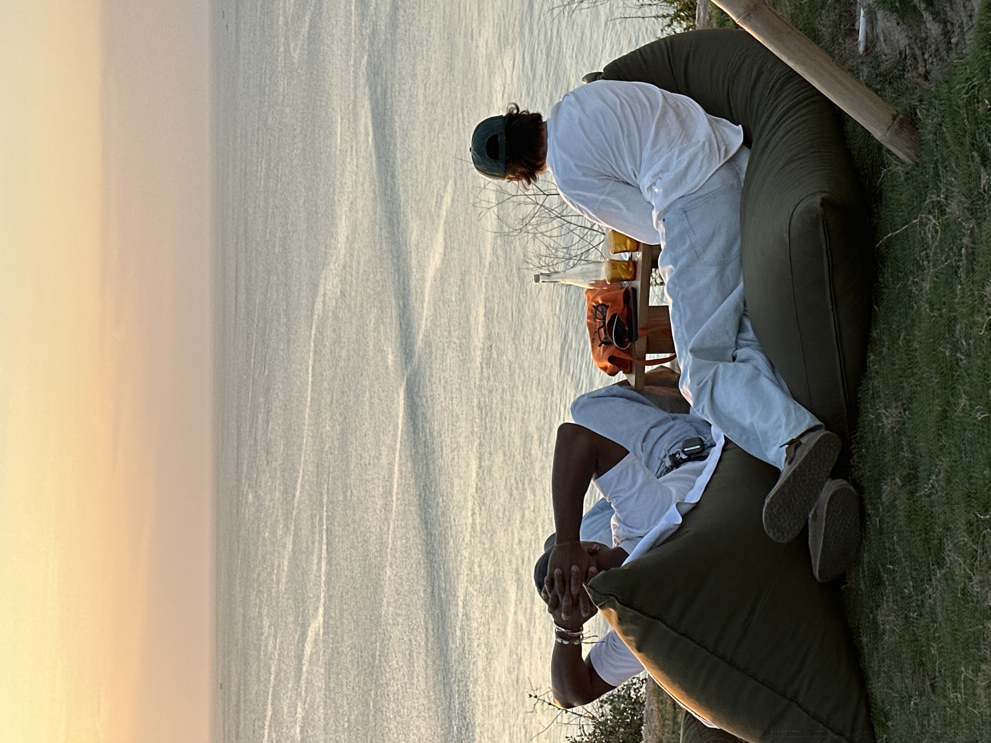 Two individuals are seated on large, olive-green beanbag chairs outdoors, overlooking a body of water at sunset. The person on the left, facing away from the camera, has their hands behind their head and is wearing a white t-shirt and light-colored pants. The person on the right, also facing away, is wearing a white long-sleeved shirt and a dark baseball cap. Between them on a wooden table are two glasses of a golden beverage, a glass bottle, and an orange bag. The setting appears to be a grassy clifftop with sparse vegetation, suggesting a location near the coast, possibly in or near Kuta Selatan, Indonesia, given the time of day and tropical environment. The water is calm with faint ripples, and the sky is a gradient of pale orange and yellow.