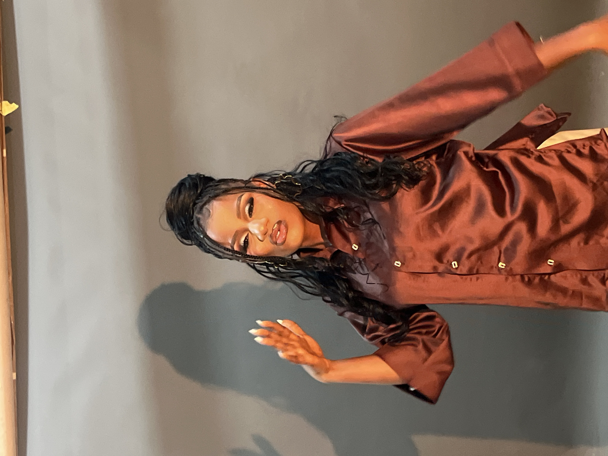 In an indoor setting with a gray backdrop, a young woman with dark, braided hair styled in a high bun and loose tendrils is captured in a medium shot. She is wearing a reddish-brown satin shirt with gold buttons and has bold makeup, including winged eyeliner and dark lipstick. Her left arm is raised and bent at the elbow, with her hand open and facing the camera, displaying long, manicured nails. A shadow of the woman is cast on the wall behind her. The location is identified as Gura Top, Nigeria.