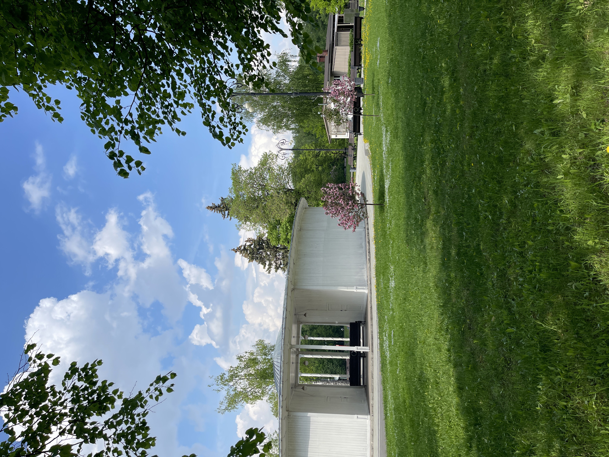 The image shows a white gazebo with benches inside, surrounded by green grass and trees, under a blue sky with white clouds. The gazebo is located in a park or garden, possibly in the city of Unknown, Unknown. There are lampposts and flowering trees visible along a path that runs adjacent to the grass area. A building with a porch is visible in the background. The image is oriented vertically.
