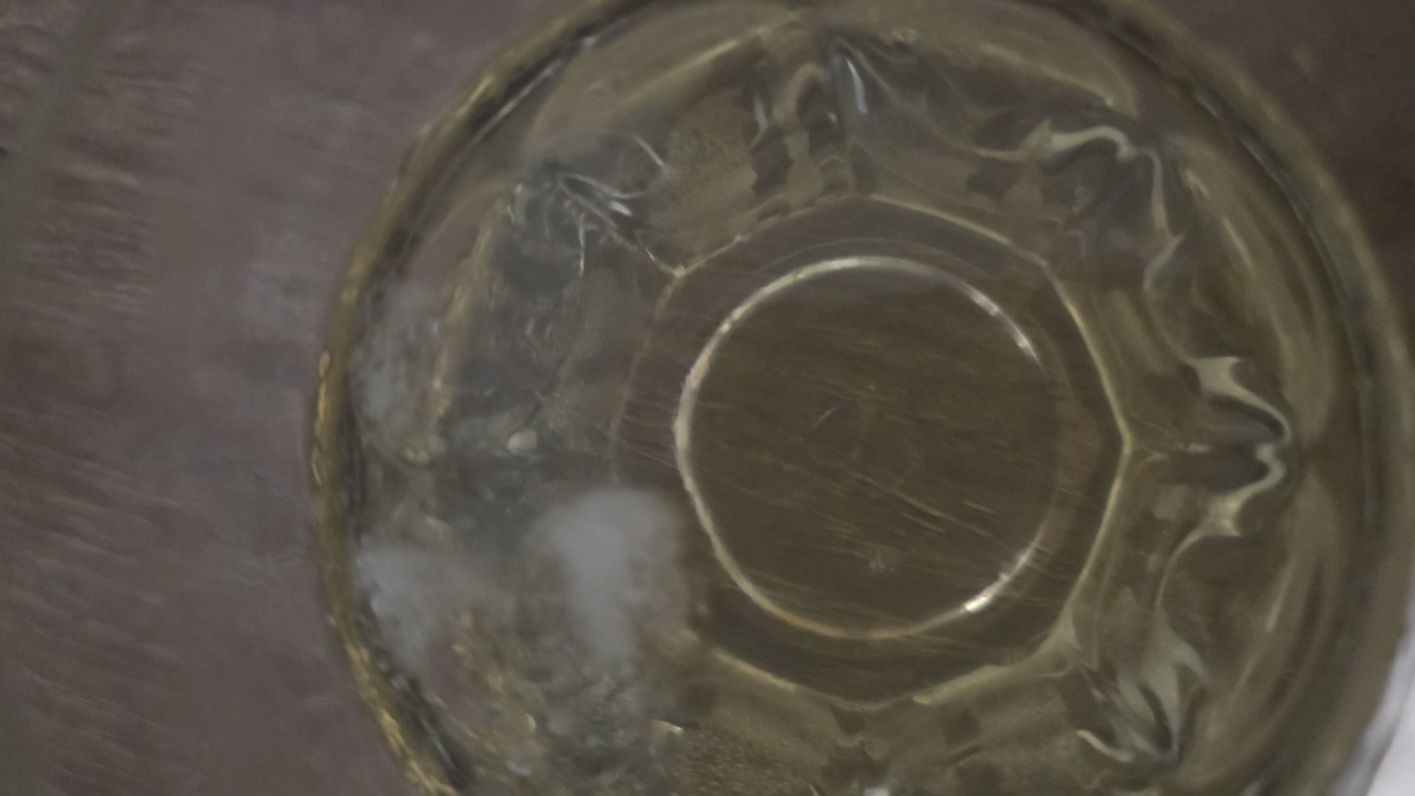 The image is a close-up, overhead view of the bottom of a clear glass or bowl, possibly containing a clear liquid. The surface the bowl is resting on appears to be a wooden or faux-wood surface. There's a white, cloudy residue or stain on the inside of the glass towards the left edge. The inner bottom of the bowl is scratched. There is no identifiable information to determine its location in the city of Blagoevgrad, Bulgaria or to identify any people, actions, or interactions.