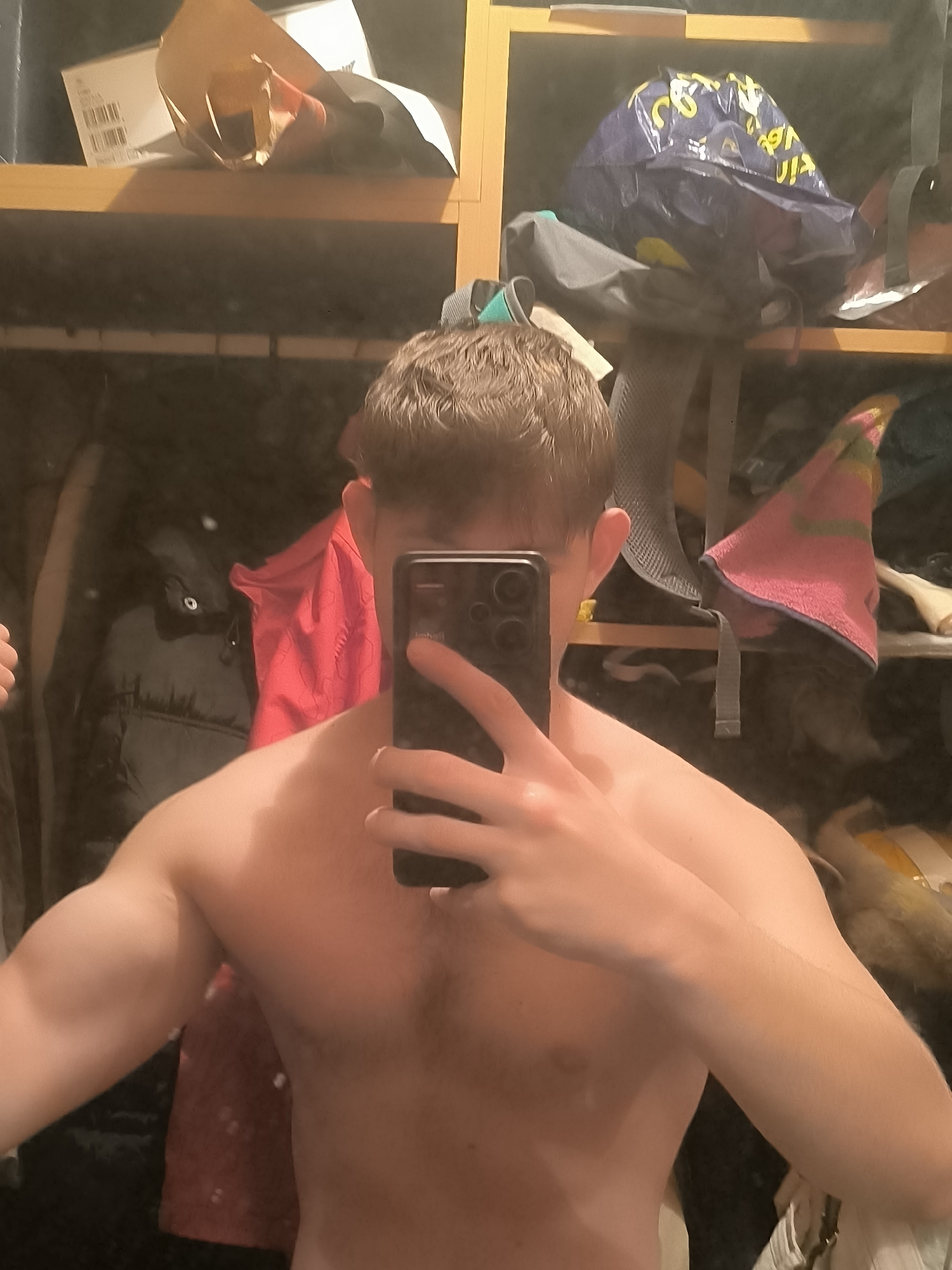 This image is a mirror selfie taken by a young man in what appears to be an indoor setting in Sofia, Bulgaria. The subject, a shirtless young man, is visible from the chest up. He has short, brown hair and appears to be in good physical shape, with noticeable muscle definition in his arms and shoulders. He holds a dark-colored smartphone in his right hand, covering most of his face and obscuring his expression. The smartphone features multiple camera lenses on its rear.
The background, reflected in the mirror, shows a cluttered shelving unit constructed from light-colored wood. On the top left shelf, a brown paper bag or box rests beside a white box that displays an unreadable barcode sticker. The top right shelf holds a blue plastic shopping bag with yellow lettering, which appears reversed due to the reflection, showing fragments like "You" and possibly "Via". Below this, a dark grey or black backpack is visible. Further down and to the right, a pink and yellow striped towel or cloth is draped or folded. On the left side of the reflection, a red garment, possibly a jacket or shirt, hangs or is draped, next to a dark puffer jacket.
The lighting in the scene is dim, suggesting an indoor environment illuminated by artificial light, possibly during the evening or night. The arrangement of personal items on the shelves points to a private residential space, such as a bedroom or dressing area within a home in Sofia, Bulgaria. The act of taking a shirtless selfie suggests a display of confidence or physical form.